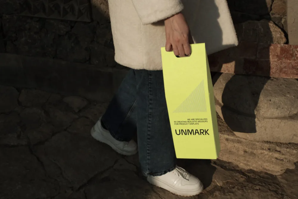 Hand holding a bright yellow vertical bag mockup with bold branding. Set in an outdoor environment with textured stone pavement and warm sunlight casting shadows.