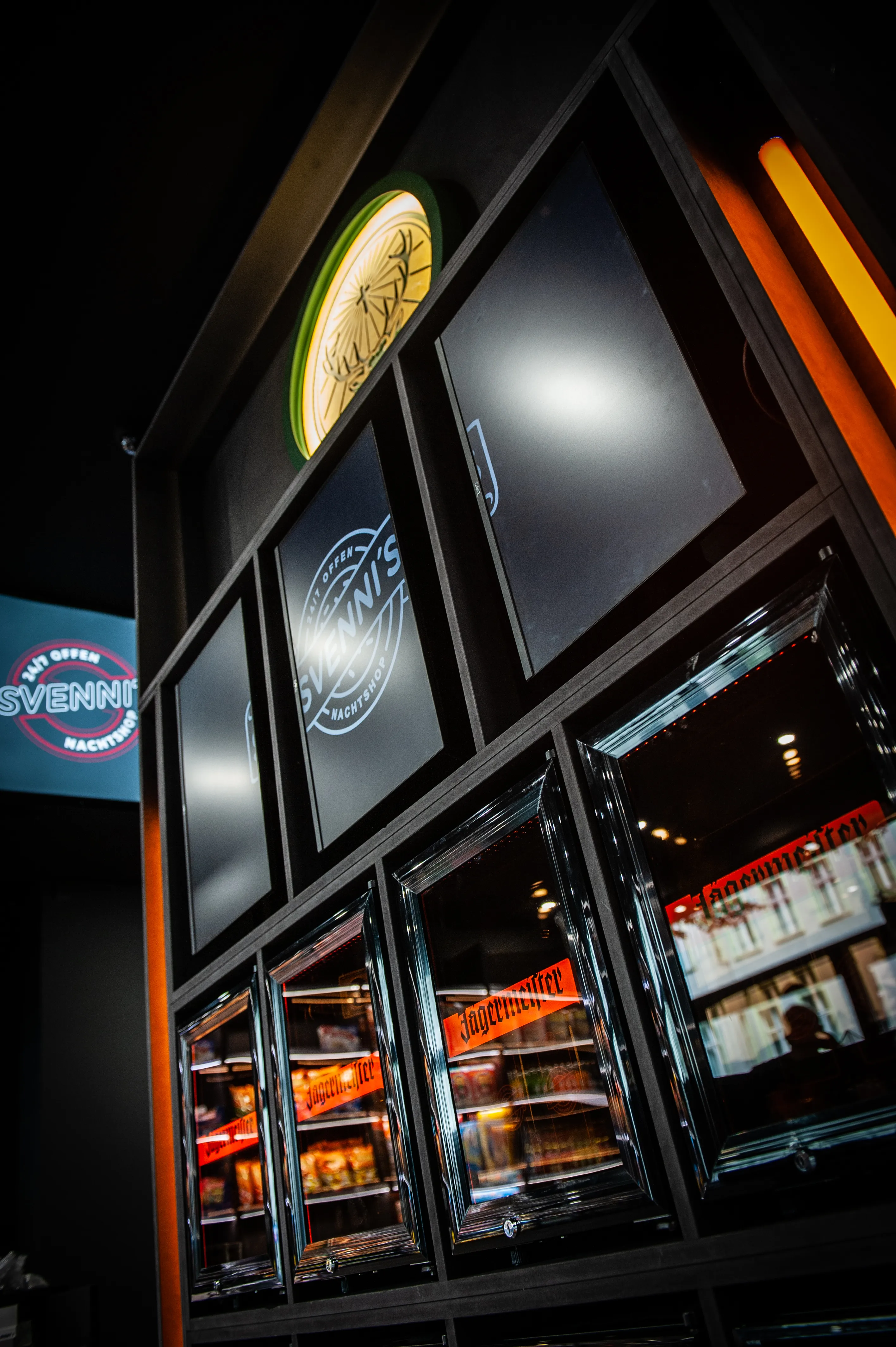 Upward view of the dark fridge wall at JÄGERMEISTER x SVENNI'S with screens, a glowing Jägermeister stag emblem and the red Svenni's kiosk sign at the side.
