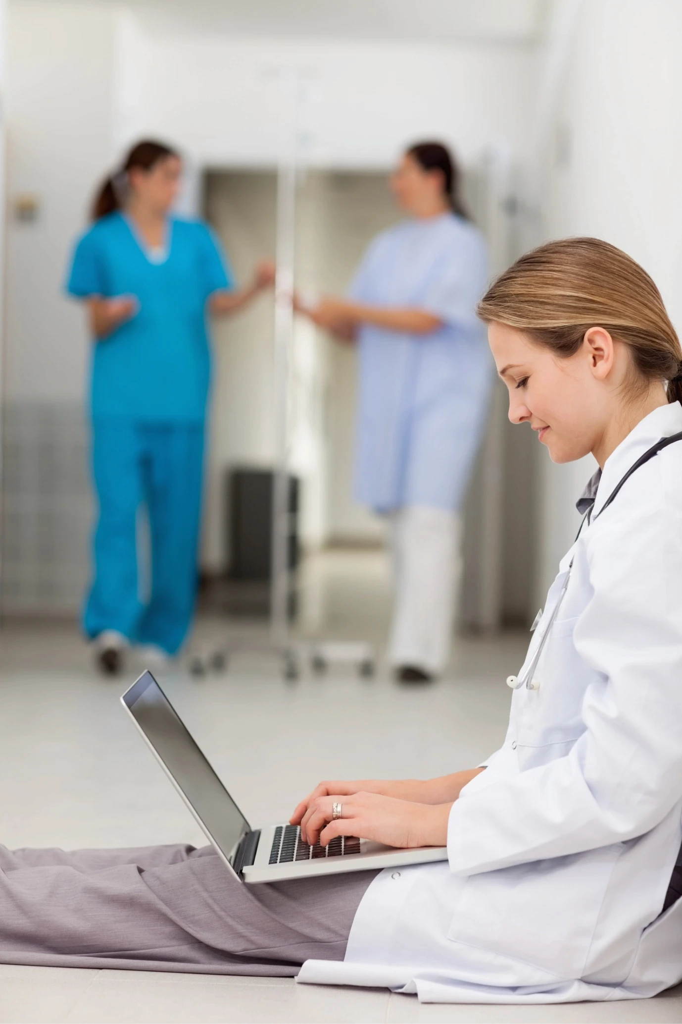 A focused female doctor in a white coat sits on the floor, working on a laptop in a hospital hallway, while two medical professionals converse in the background, highlighting a healthcare setting.