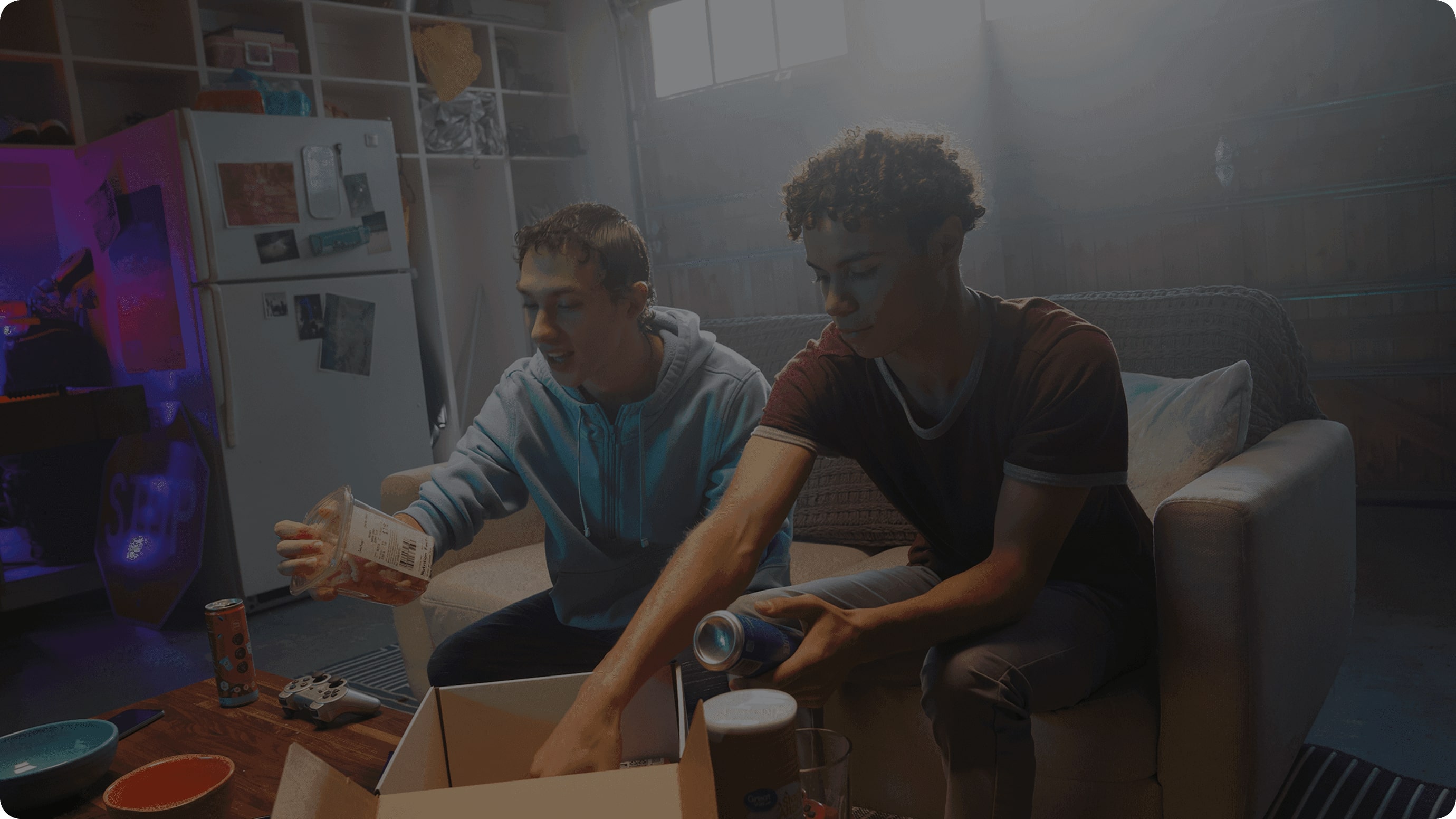 Two people sit on a couch in a dimly lit room, unpacking items from a cardboard box, with a refrigerator and shelves visible in the background.