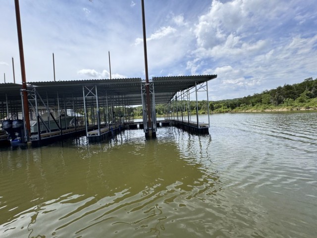 Covered boat docks with a metal roof extend over a serene lake, surrounded by lush greenery and a partially cloudy sky.