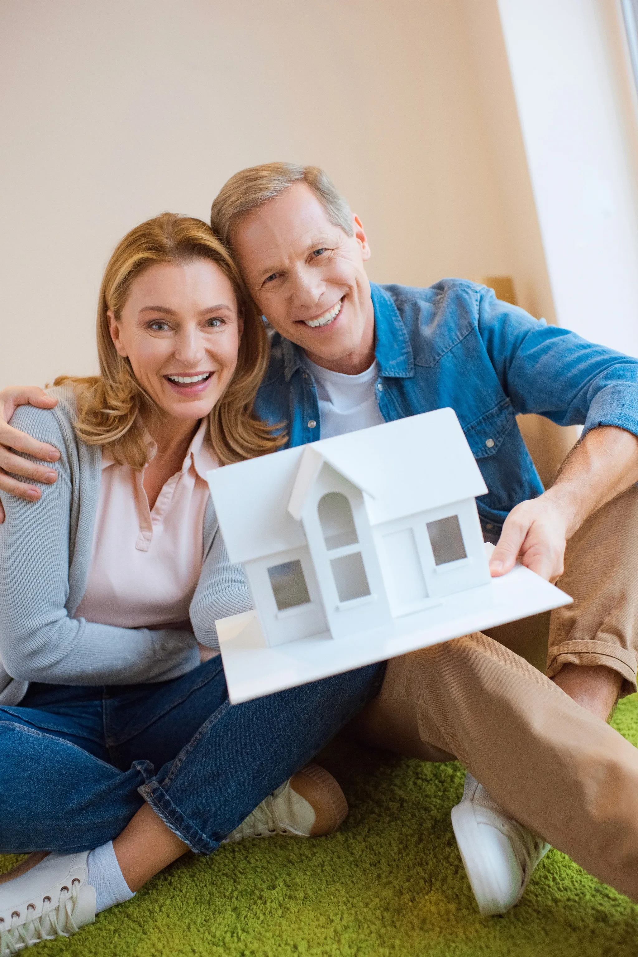 A smiling couple sits together on a green carpet holding a white architectural model of a house.