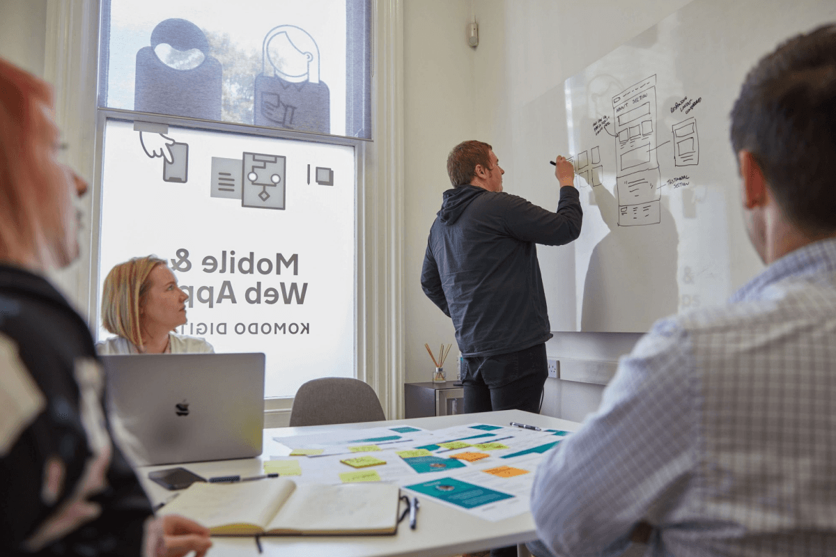 A workshop at the Komodo studio. There are post it notes on the table and one of the designers is sketching wireframes on a whiteboard