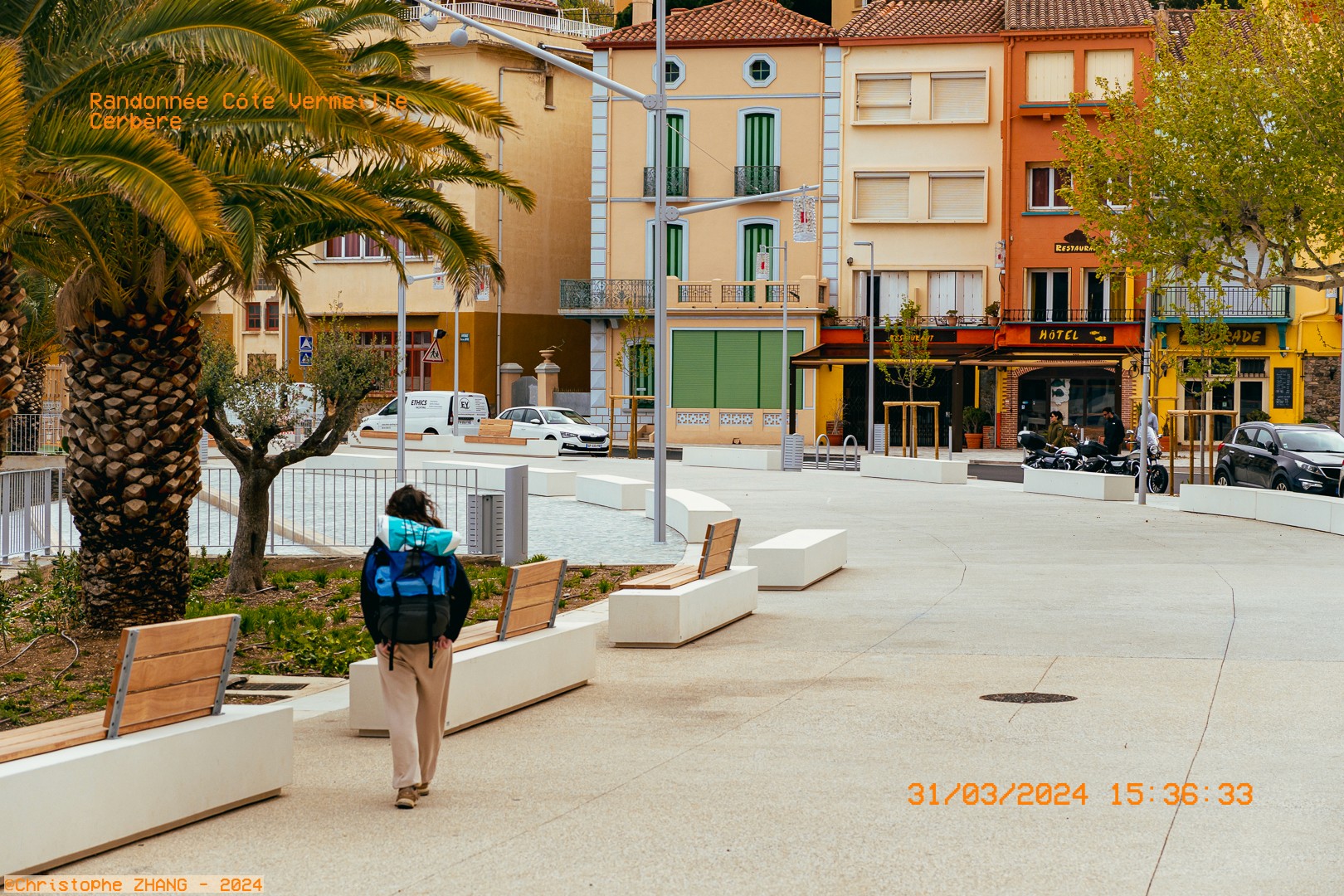 Personne en randonnée traversant une place urbaine bordée de palmiers à Cerbère.