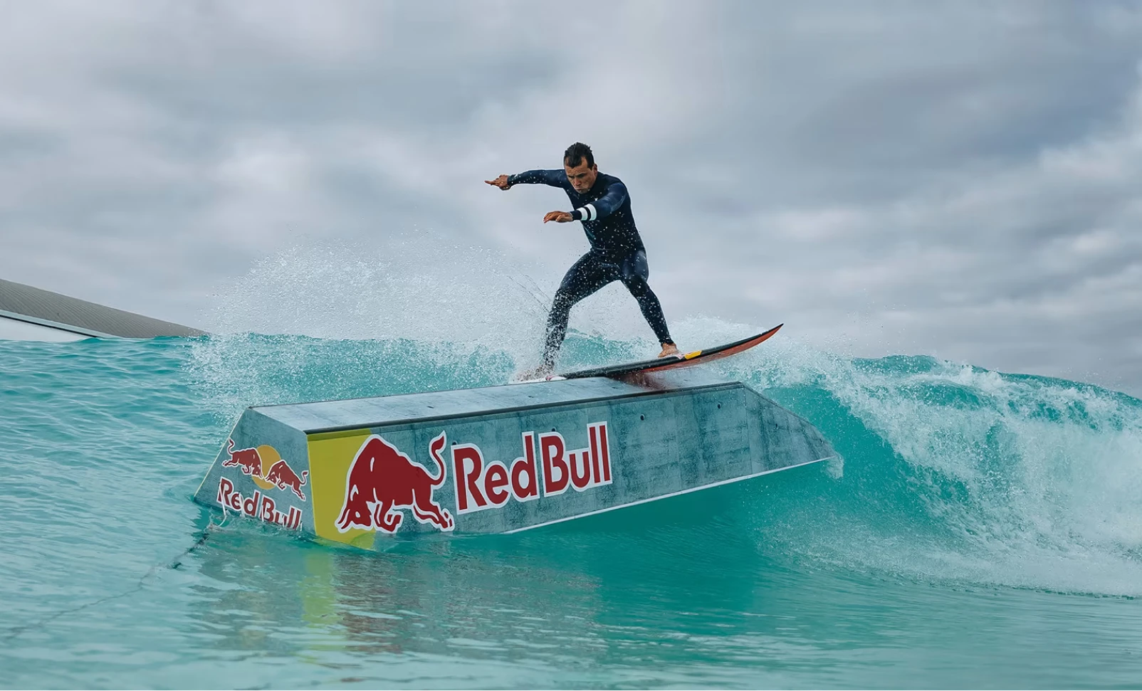 A surfer in a wetsuit skillfully rides a wave, performing a trick on a ramp with Red Bull logos, against a backdrop of a turbulent ocean and cloudy sky.