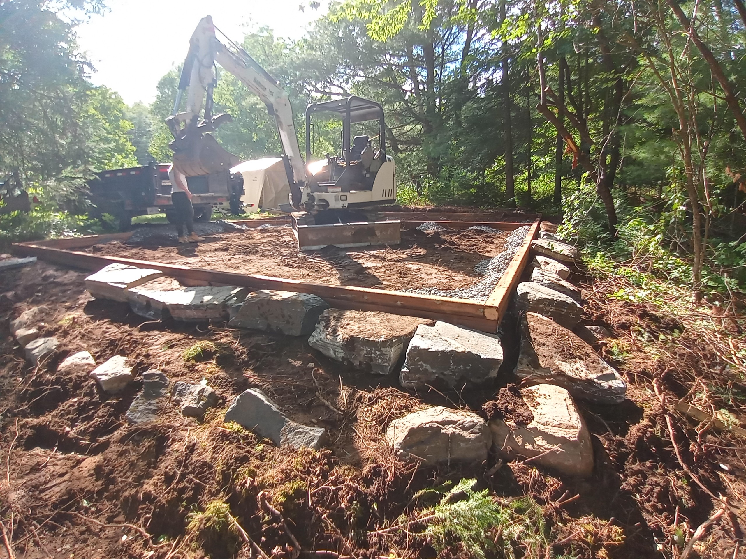 Mini excavator preparing a compacted aggregate base for a portable storage shed on a wooded cottage slope.