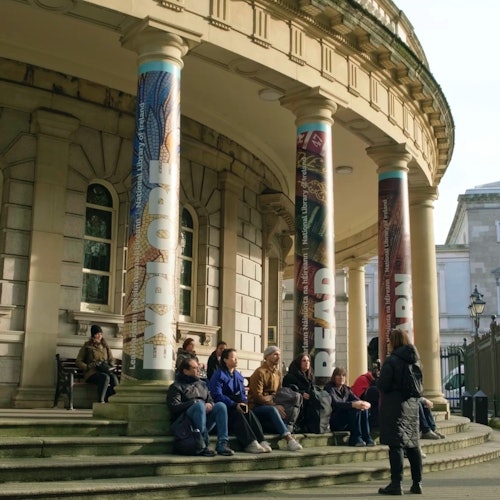 People sitting and talking on the steps of a building with large columns featuring the words "EXPLORE," "READ," and "LEARN."