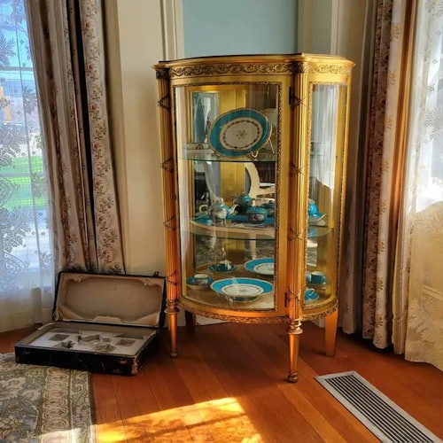 Una vetrina con rifiniture dorate contenente vari piatti blu e bianchi accanto a una valigia aperta sul pavimento in legno.