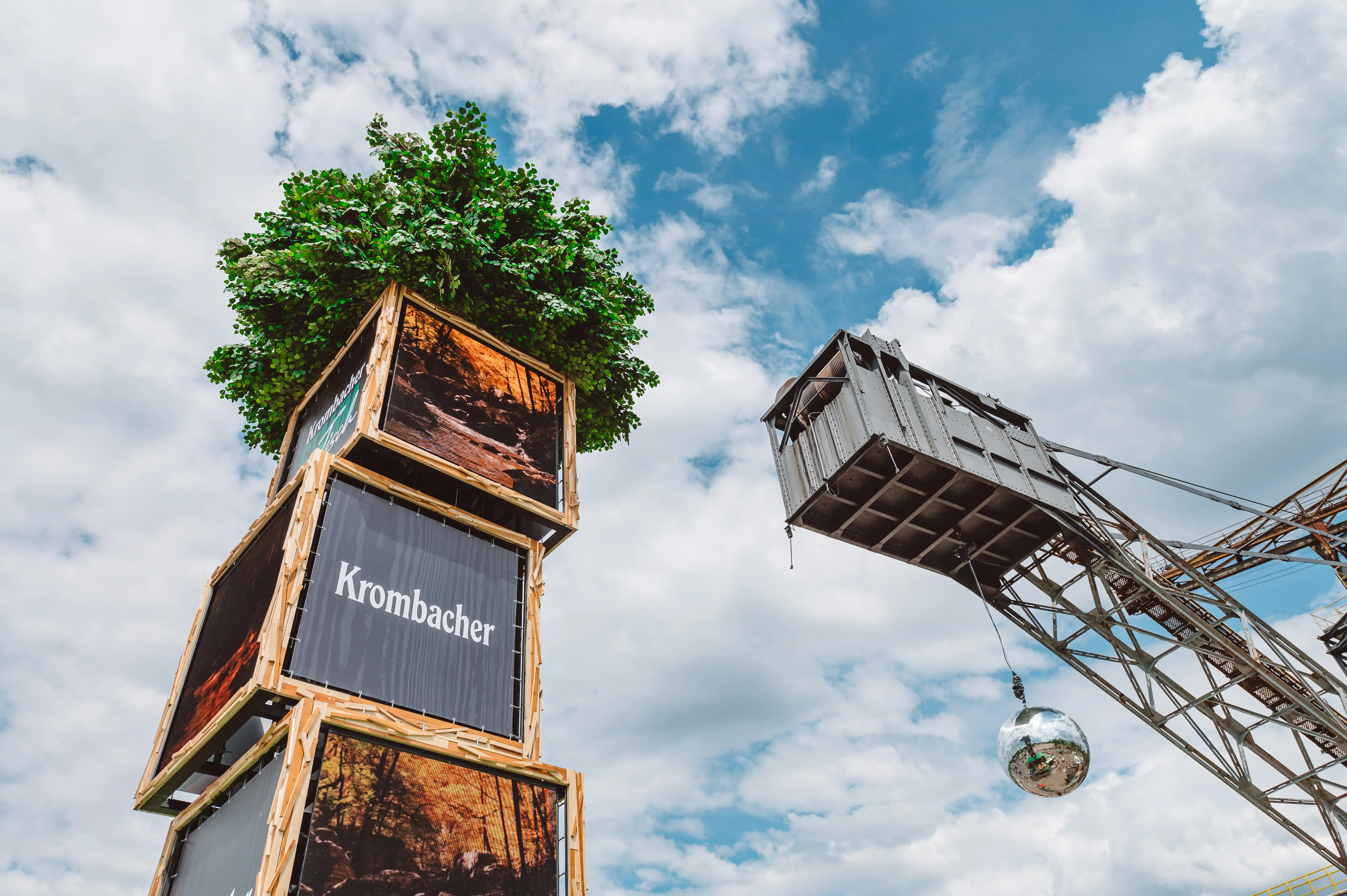Detail of the KROMBACHER crate tower with photo prints and logo panels, topped by a green tree, next to a crane arm with disco ball.