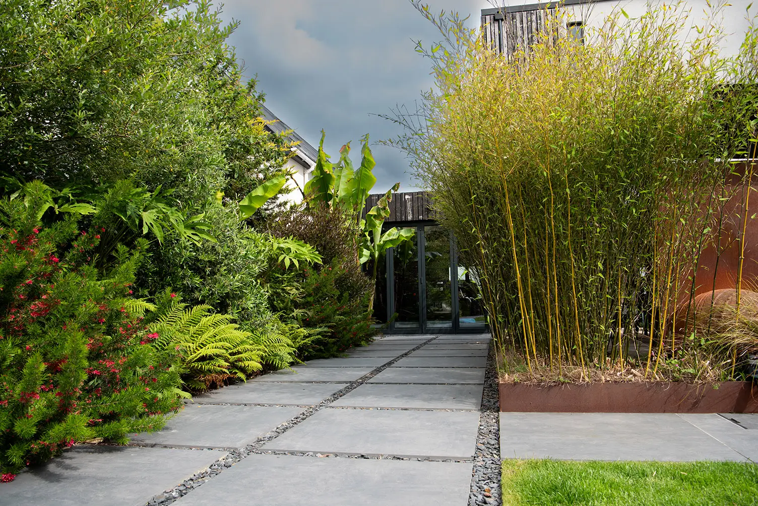 A pathway lined with lush greenery and tall bamboo, leading into a serene garden space under a cloudy sky.