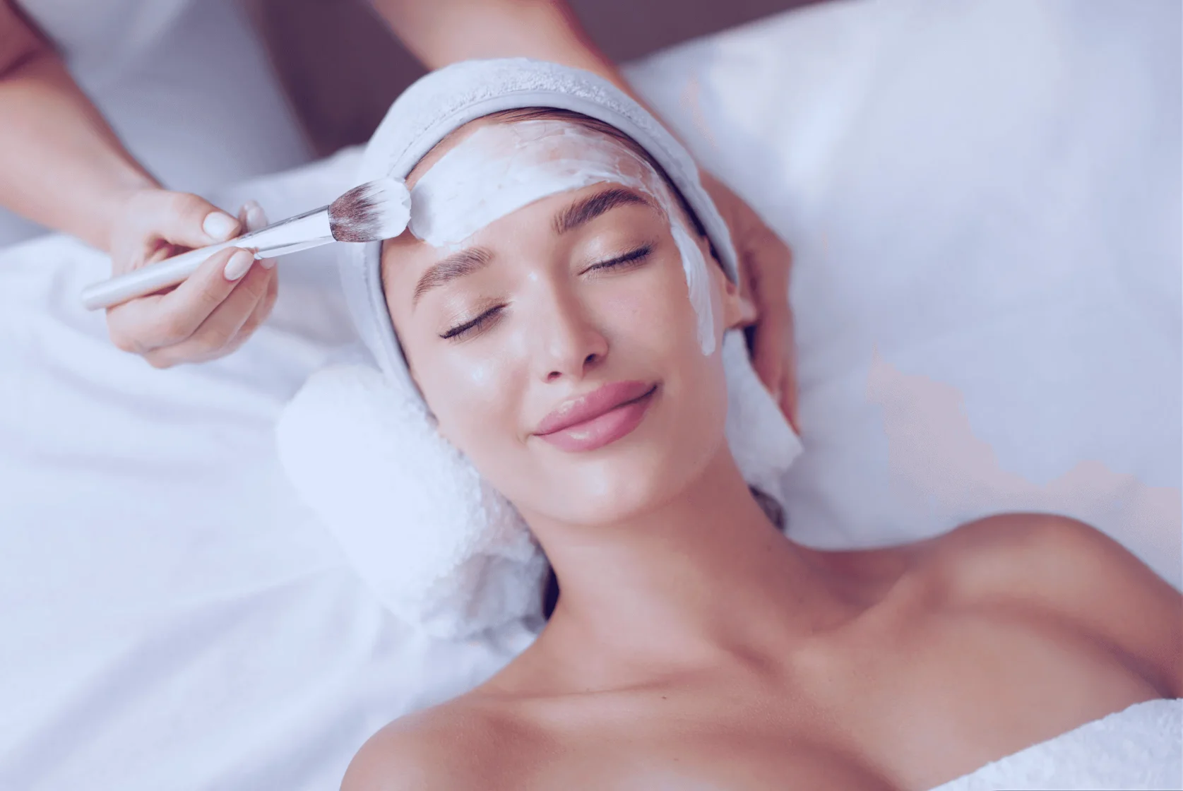 A woman enjoys a relaxing facial treatment with a cream mask applied by a beautician, lying comfortably with her head wrapped in a towel.
