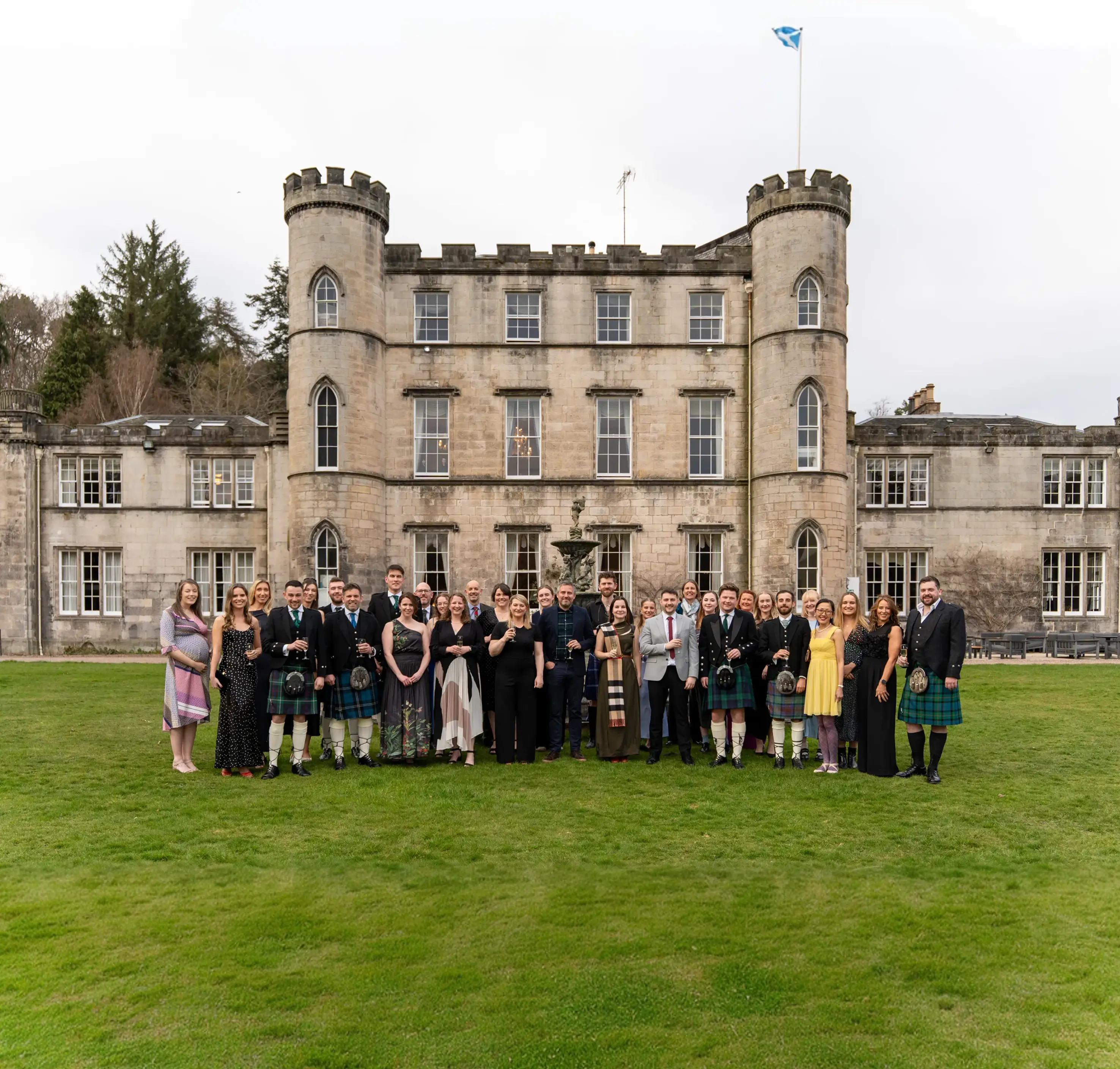 The Planning Potential team — approximately 30 employees — gathered on the lawn in front of a grand Scottish castle, flying the Saltire flag. Several team members are dressed in traditional Scottish kilts, while others wear formal evening attire, reflecting a celebratory company event.