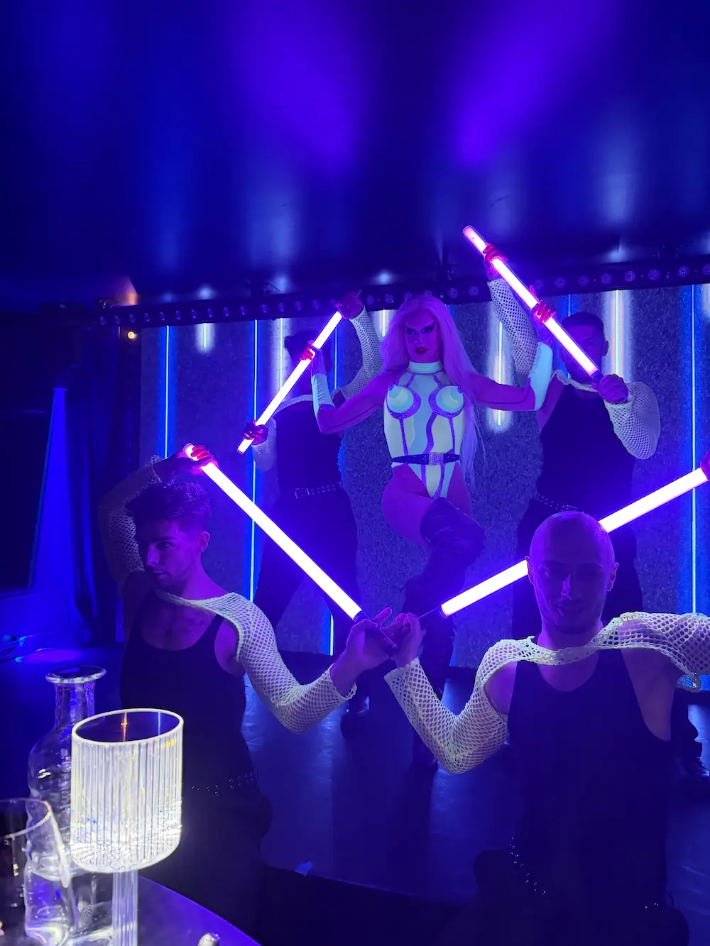 A dynamic stage shot of a central drag performer in a patterned white jumpsuit and blonde hair holding purple glowing neon sticks, flanked by two backup dancers