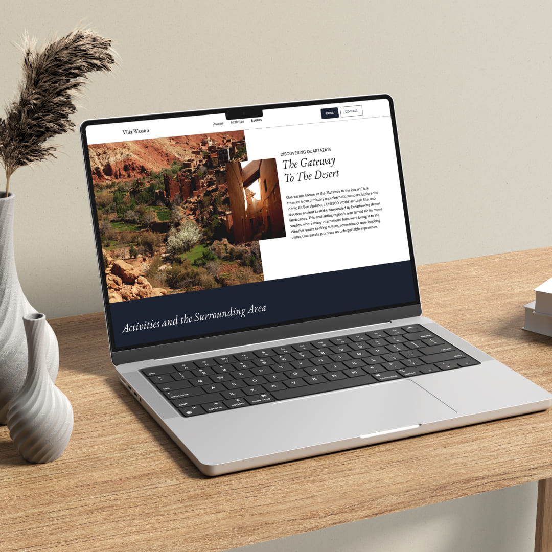 Mise en situation (mockup) du site web de Villa Wassim Marrakech, affiché sur un ordinateur portable MacBook Pro sur un bureau en bois. La page présente la section 'Activités et les alentours', avec un grand visuel de la région de Ouarzazate (paysage désertique, casbah) et le titre 'The Gateway To The Desert'. La mise en page est propre, minimaliste et haut de gamme, utilisant les polices Serif et Sans Serif de la charte graphique. Cette image démontre la qualité de l'expérience utilisateur (UX) et le design épuré réalisé par Nux-Studio pour le site web de l'hôtel de luxe Villa Wassim, facilitant la découverte des activités et du voyage.