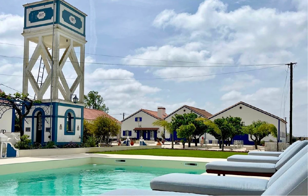 Sunny scene of a serene hotel exterior with a turquoise pool in the foreground. A tall white tower, dated 1877, stands beside white buildings with red roofs.