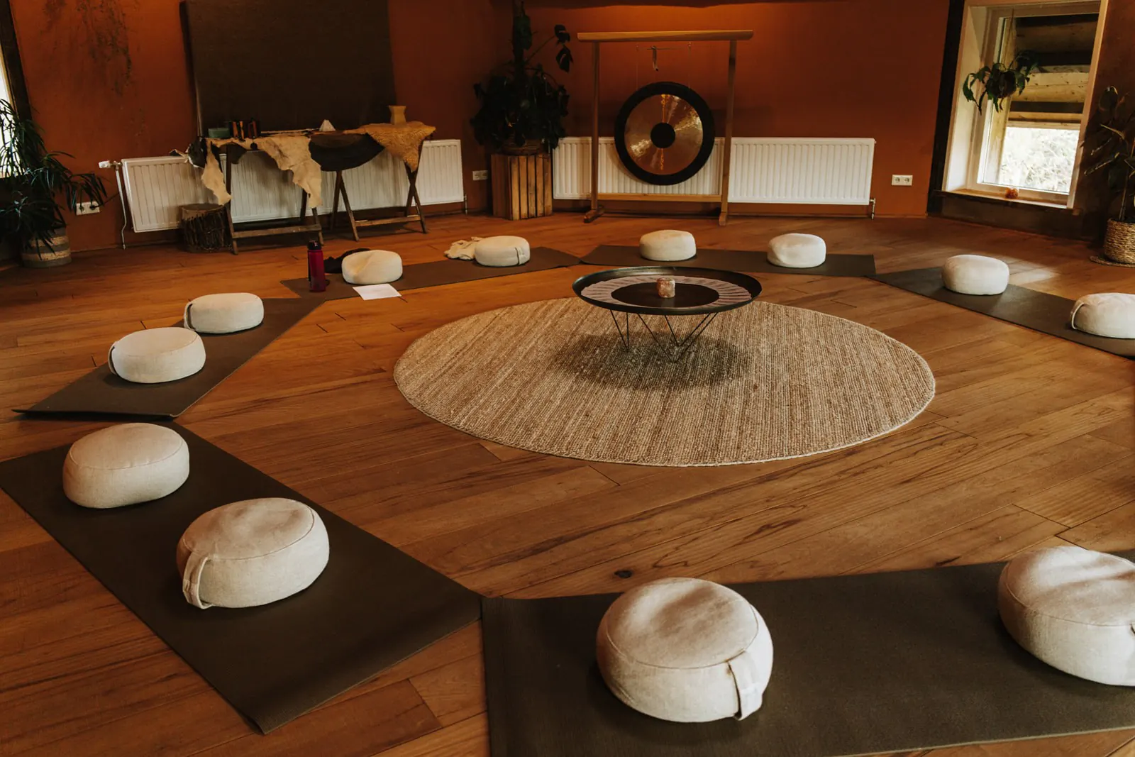 Een lege ruimte met houten vloer en witte zitkussens op matjes in een kring om een rond kleed, achterin een gong