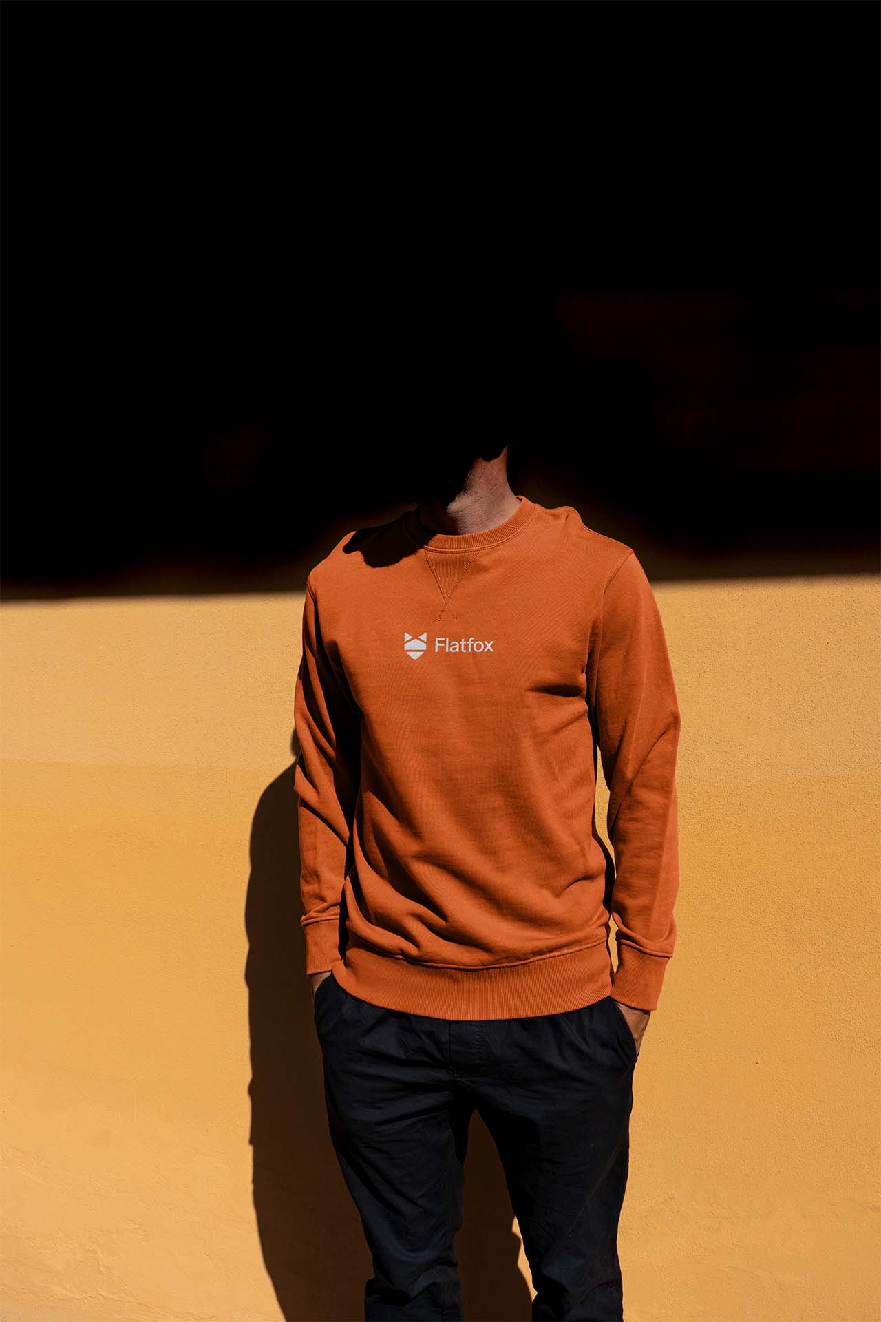 A person wearing a rust-orange crewneck sweatshirt featuring a minimalist white Flatfox logo. This piece of corporate identity collateral demonstrates how the brand's visual language translates into premium, modern merchandise for the tech sector.