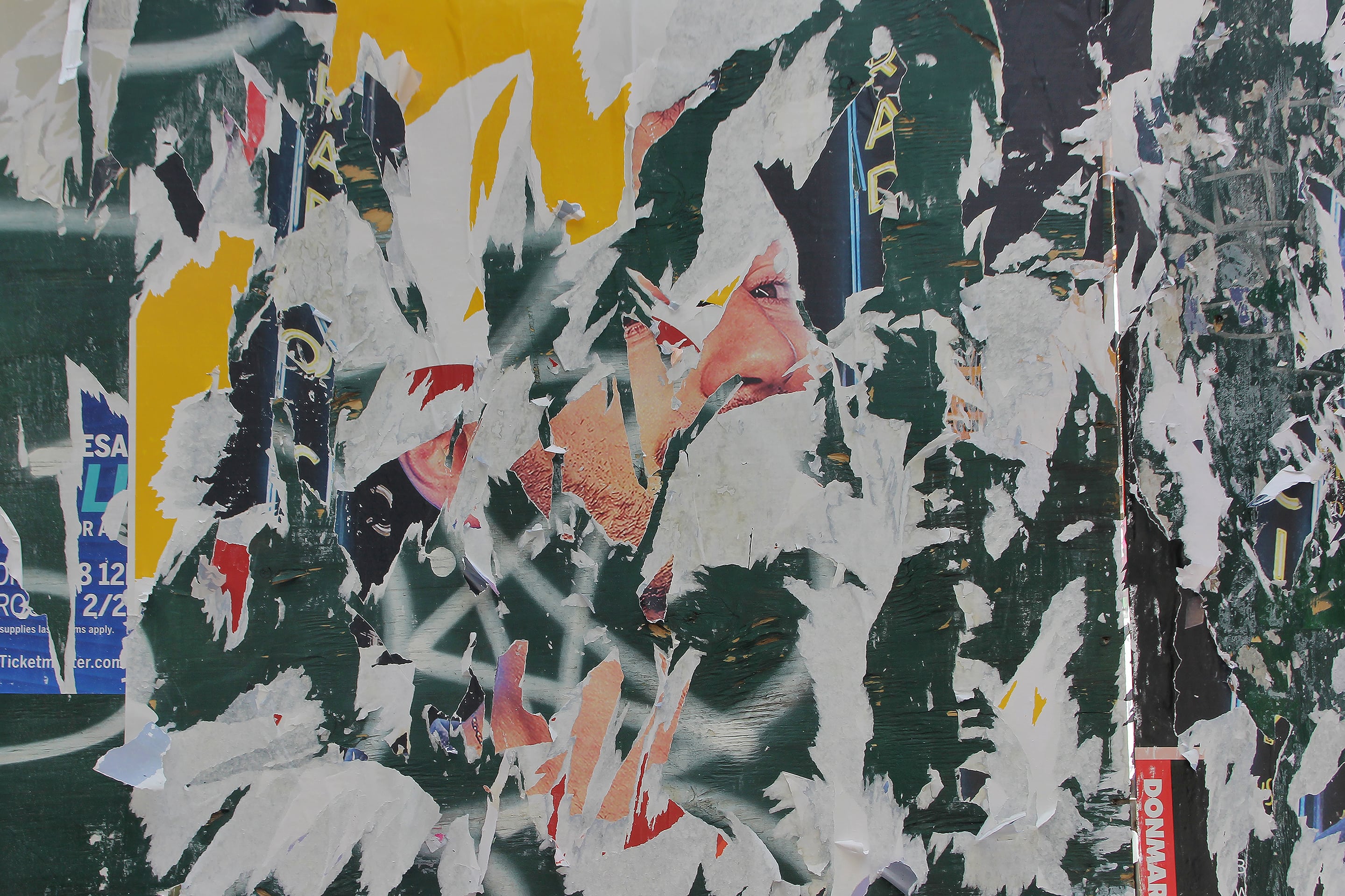 Photograph of a torn poster wall revealing fragments of a man’s face beneath layers of green, white, and yellow paper. The jagged textures and overlapping imagery create a fragmented portrait that suggests erosion, concealment, and transformation.