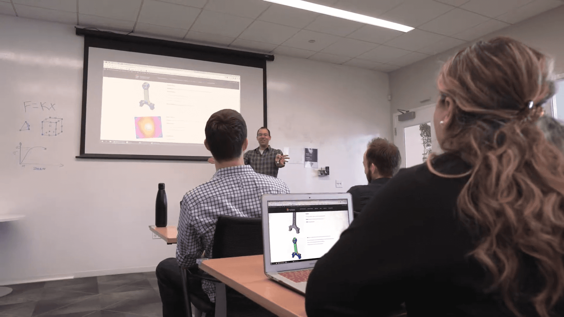 A classroom scene with students sitting at desks while a professor presents on a screen, showing a technical diagram or 3D model