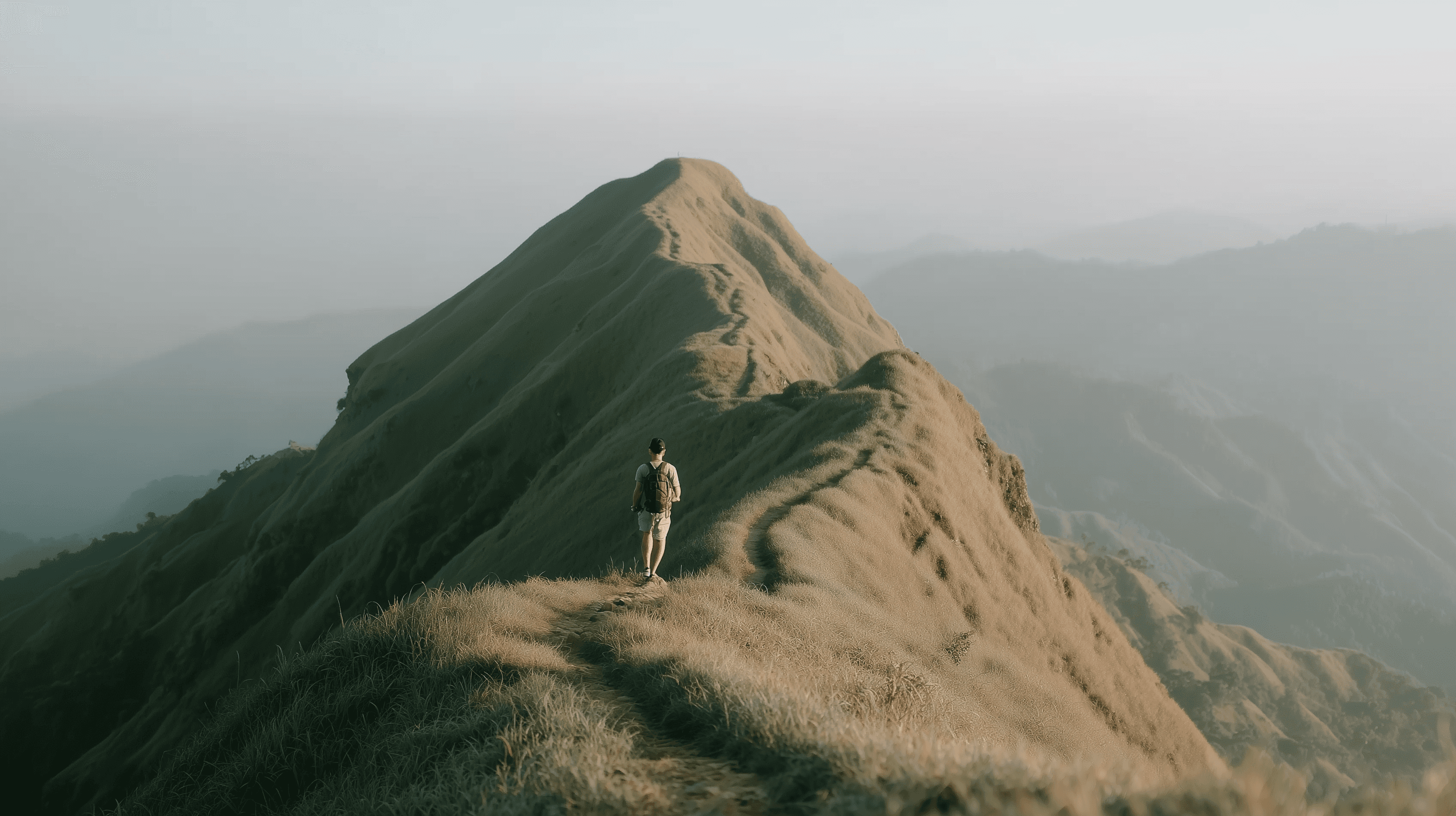 Hiker walking along a narrow mountain ridge at sunrise, surrounded by misty rolling hills.