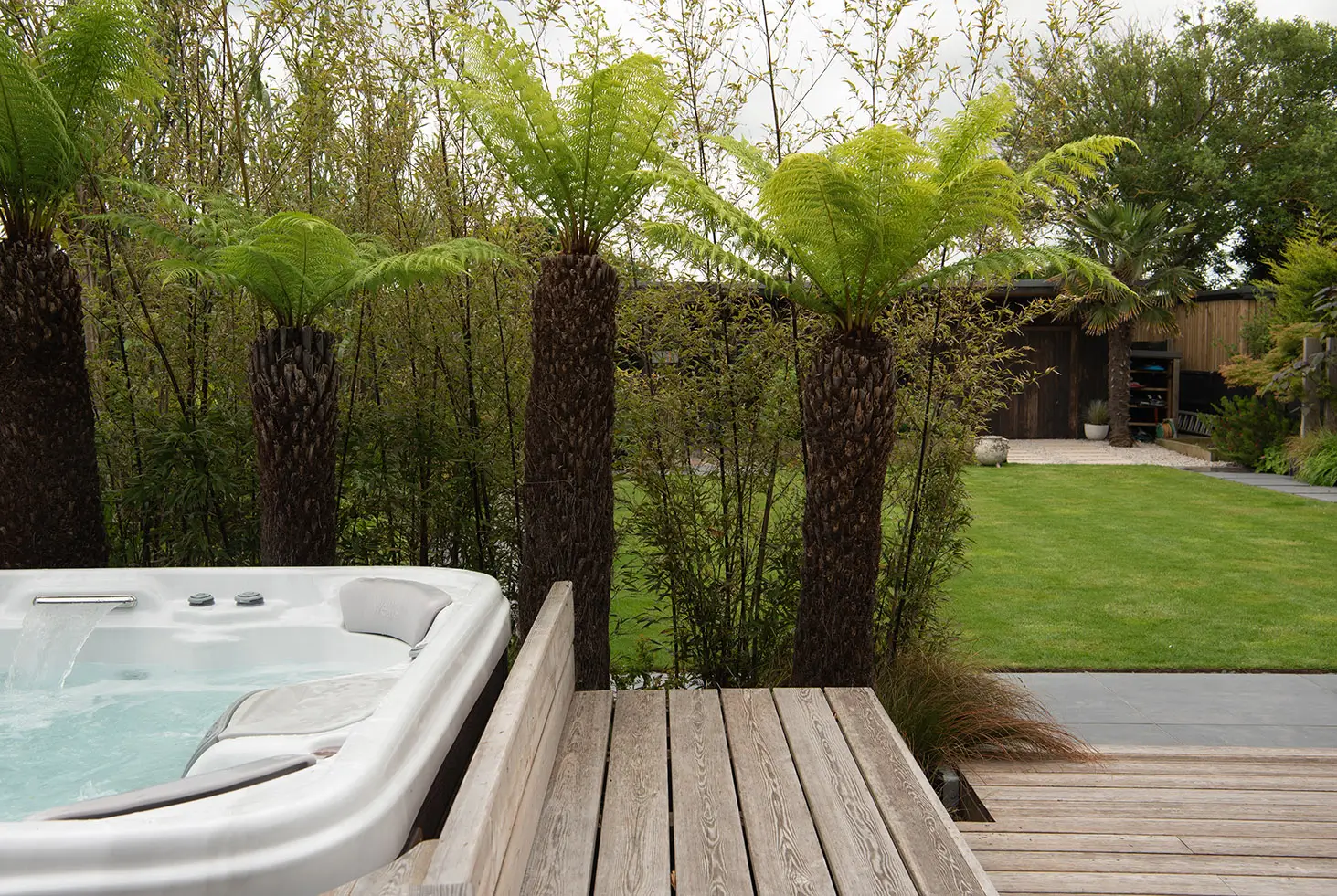 A wooden deck with a hot tub and lush green plants in the background, set in a peaceful outdoor environment.