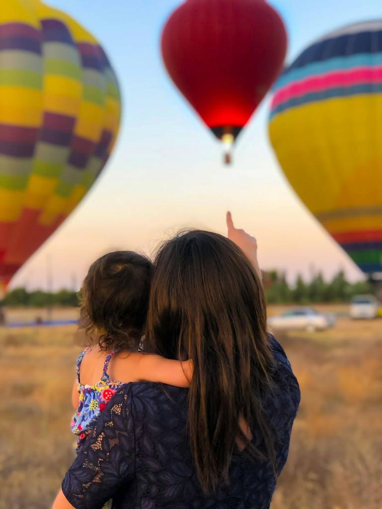 A mother and her young child admire colorful hot air balloons rising into the sky, capturing a moment of wonder and bonding at a hot air balloon festival.