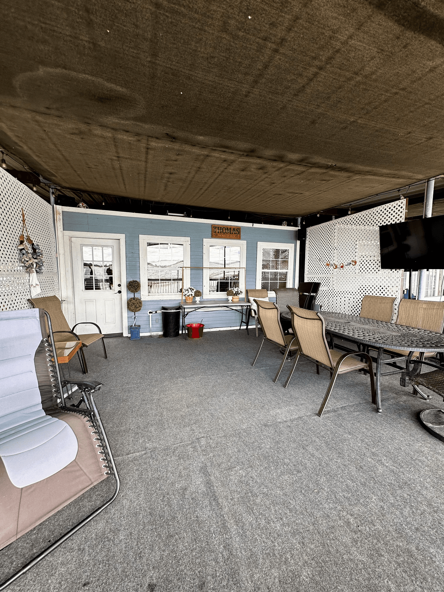 A spacious outdoor patio features a covered seating area with multiple chairs around a large table, set against a backdrop of white lattice walls, a blue building, and potted plants, creating a relaxed and inviting atmosphere.