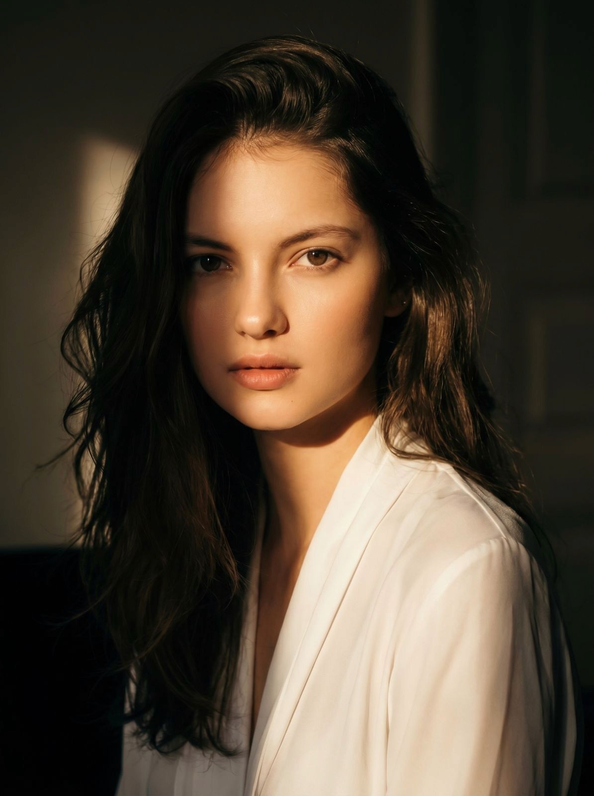 A woman with long dark hair is gazing softly at the camera, illuminated by warm, natural light.