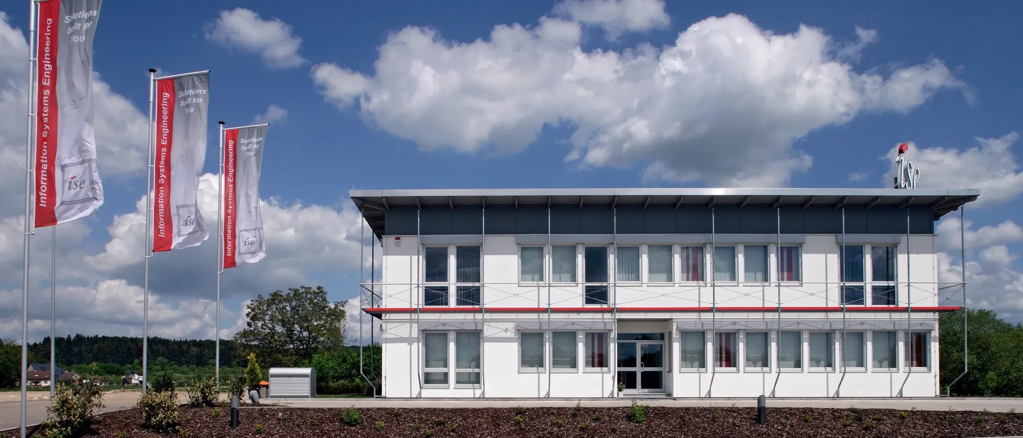 Modernes Bürogebäude mit Fahnenmast im Vordergrund und blauem Himmel mit Wolken.