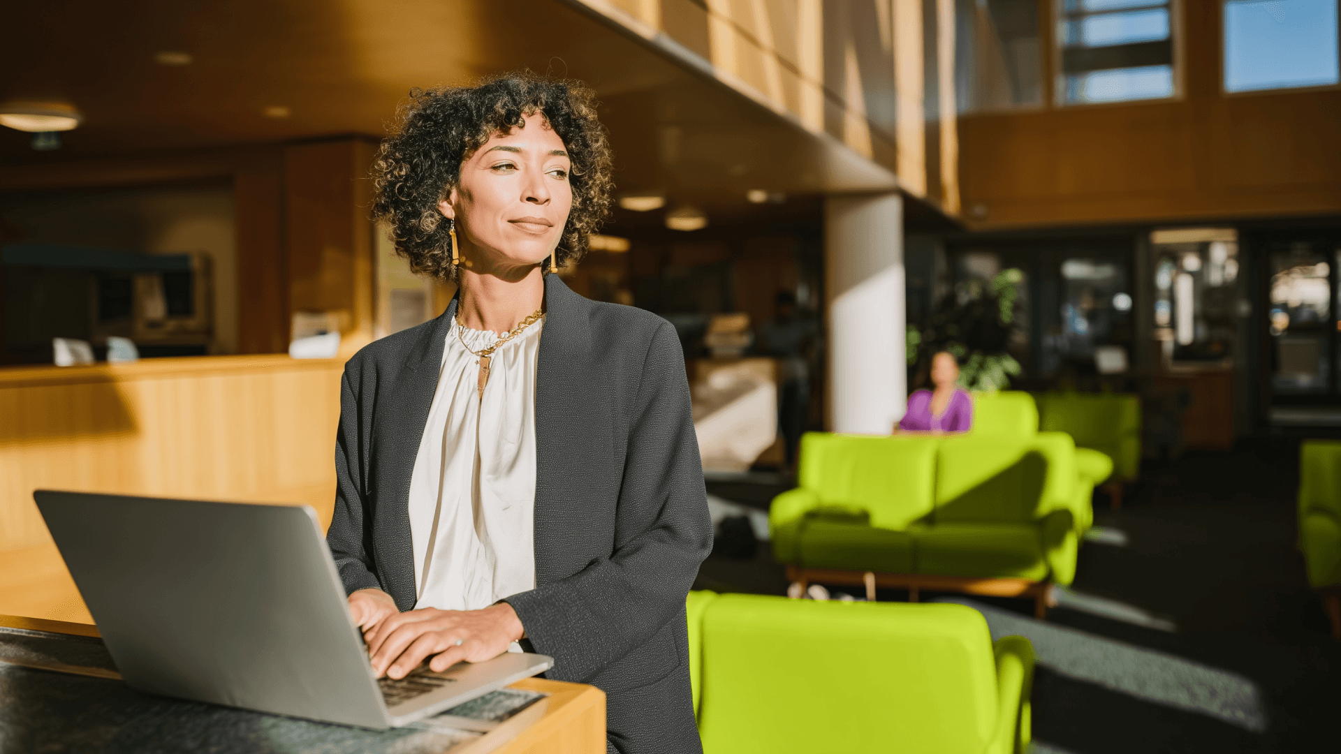 Foto de uma mulher com cabelos cacheados e blazer cinza, em pé na recepção de uma clínica bem iluminada com sofás verdes ao fundo. Ela está usando um notebook na Recepção da Clínica. A imagem simboliza a preparação antes de receber um paciente com o auxílio da Tivita.