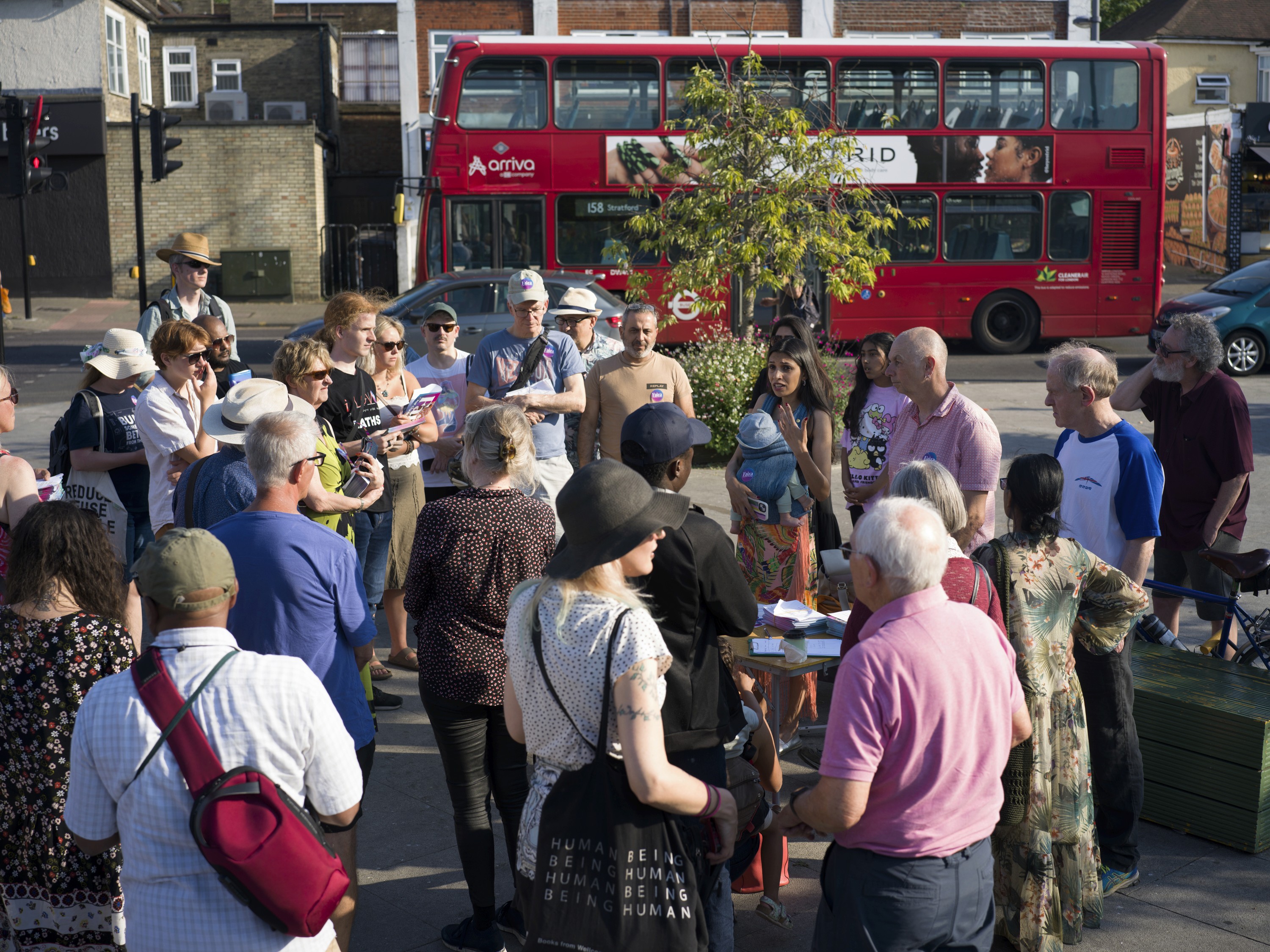 Group of people outside at a community stall listening to a speaker. A 158 bus going past in the background.