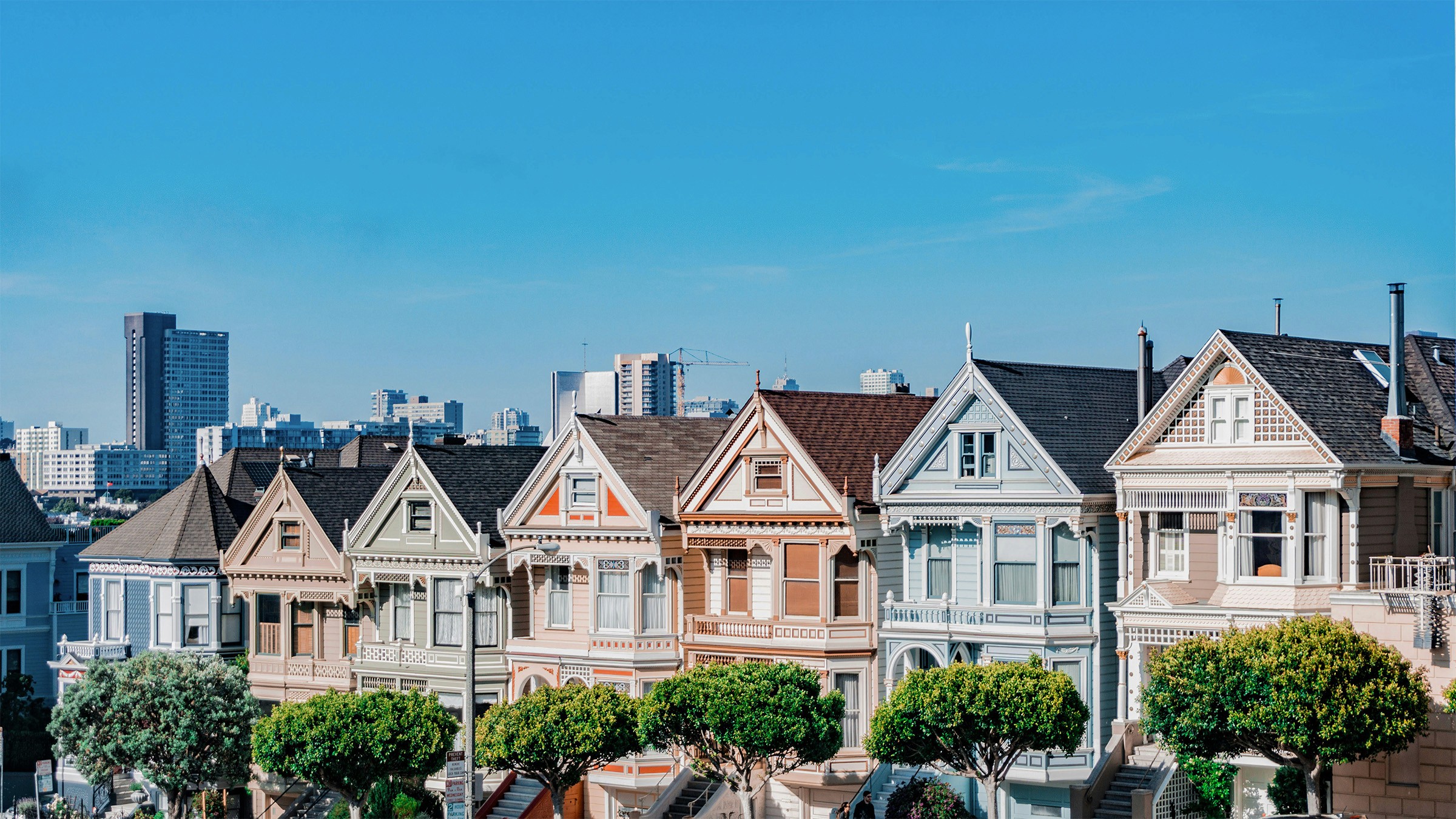 Colorful Victorian-style houses, known as the 'Painted Ladies,' line a sunny street with a city skyline and high-rise buildings in the background under a clear blue sky