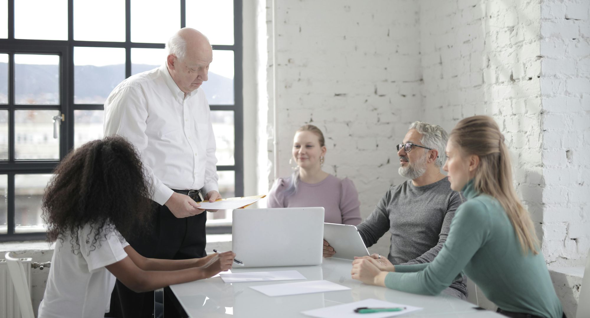 pessoas sentadas à uma mesa com notebooks e uma de pé, são três mulheres jovens e dois homens de meia idade