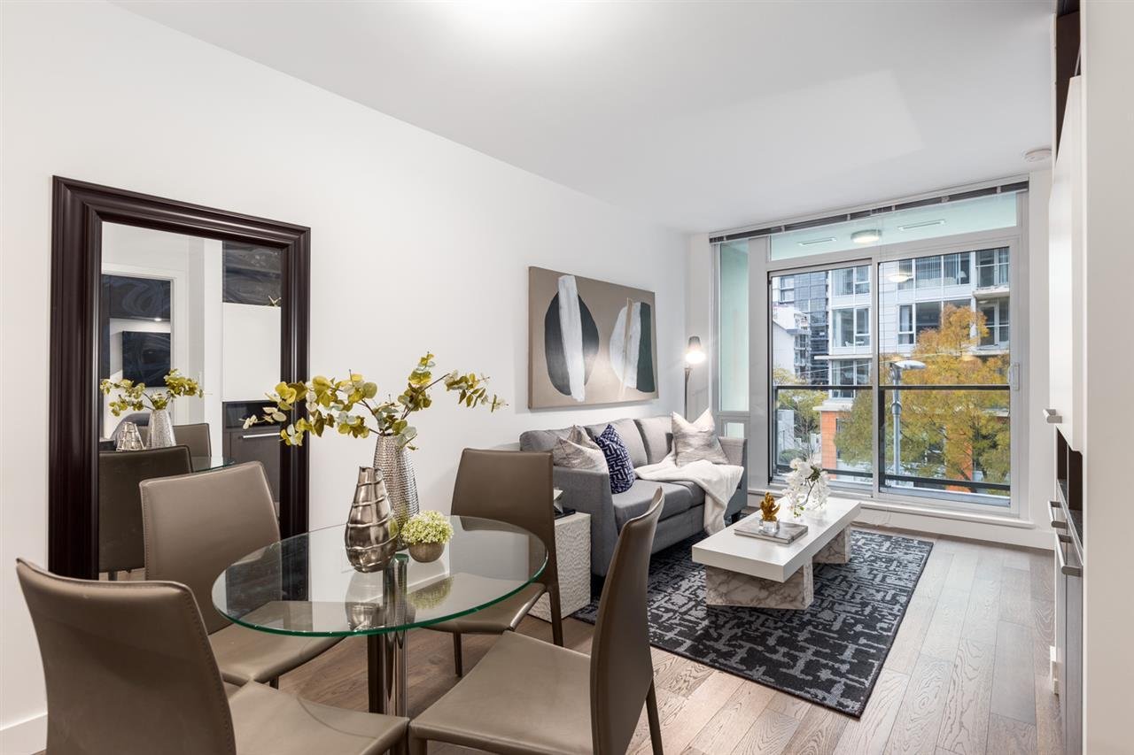 Modern living room with a glass table, chairs, artwork on the walls, and large windows allowing natural light.