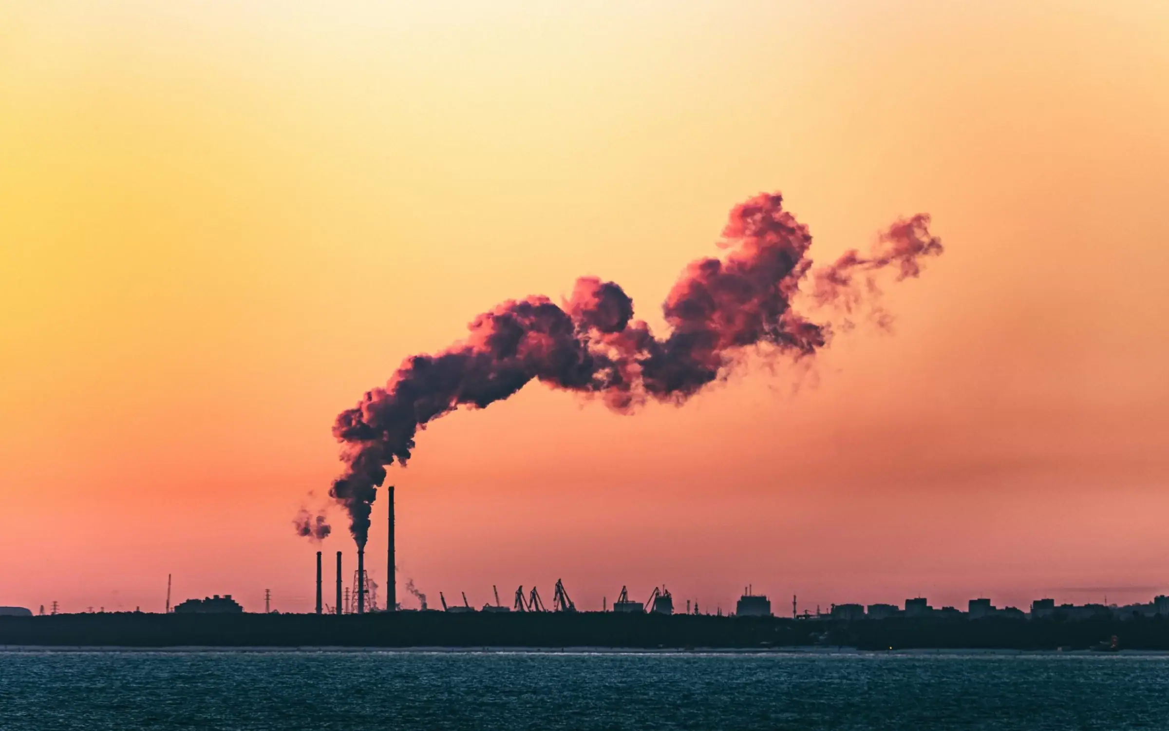 Industrieanlage an der Küste mit dichtem Rauch bei Sonnenuntergang – Symbol für Emissionsquellen und Emissionsfaktoren-Datenbanken