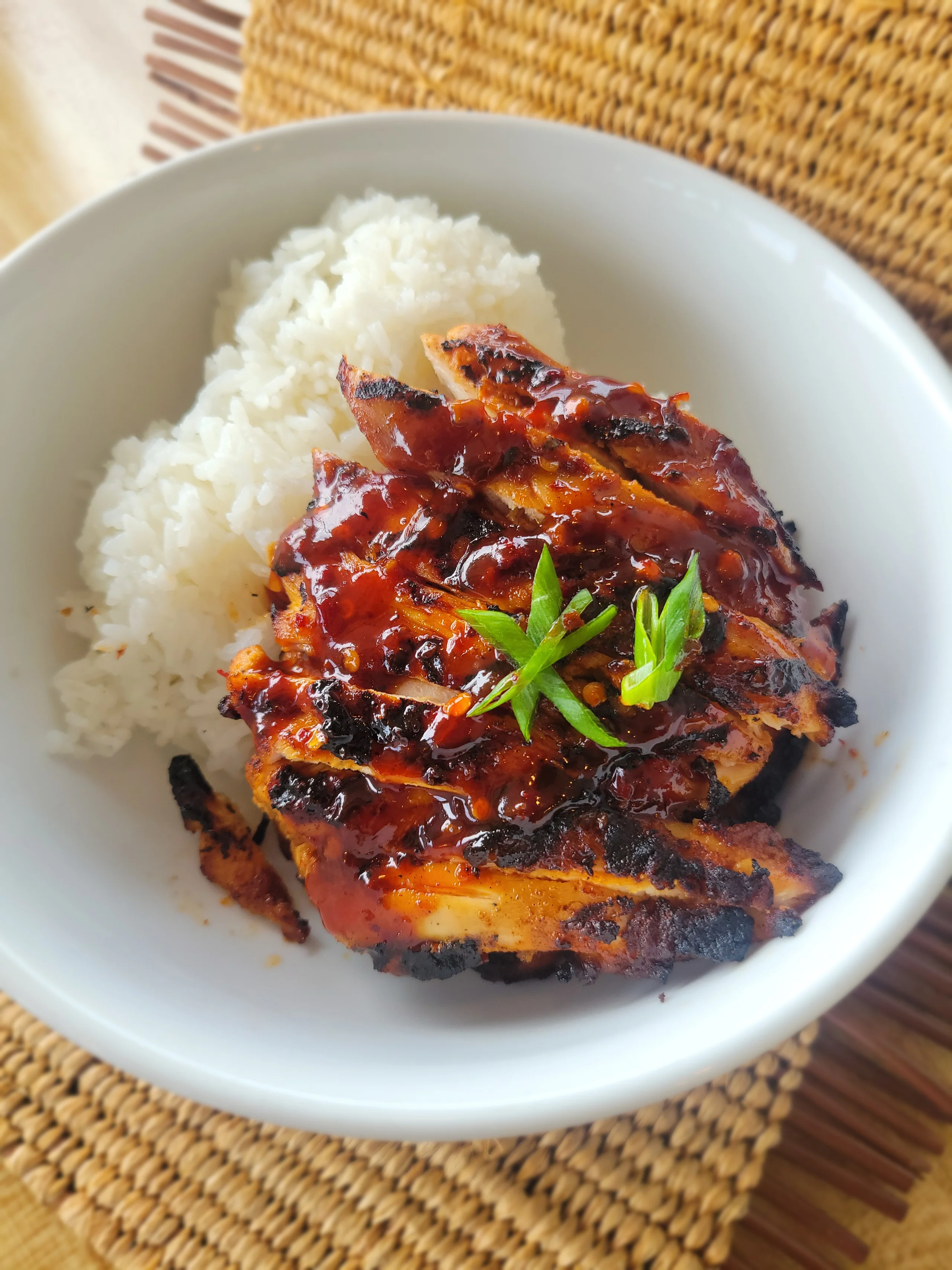 A white bowl filled with sliced, char-broiled chicken thighs glazed in a vibrant red spicy sauce and garnished with green onions, served next to a portion of white rice.