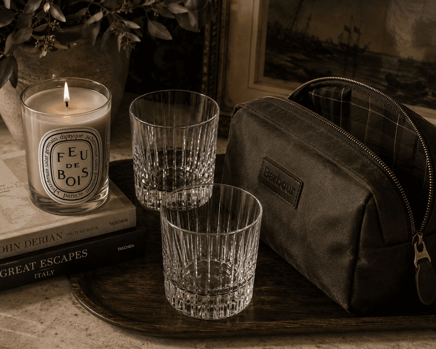 A moody still life featuring a lit Diptyque "Feu de Bois" scented candle resting on stacked Taschen art books next to two clear cut-crystal tumbler glasses. Alongside them on a dark wooden tray sits an open Barbour olive green waxed canvas toiletry bag showing its plaid fabric lining, with framed artwork softly lit in the background.