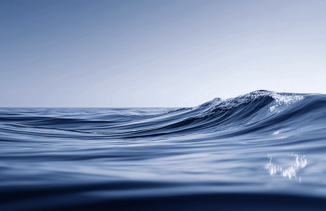 Vista em close da superfície da água do mar, mostrando pequenas ondas suaves e texturizadas em tons de azul profundo. Ao fundo, uma linha do horizonte nítida separa o oceano de um céu azul claro e sem nuvens