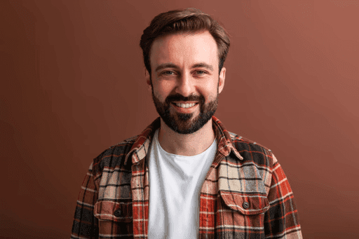 Young man with beard smiling happily in plaid shirt against brown background.