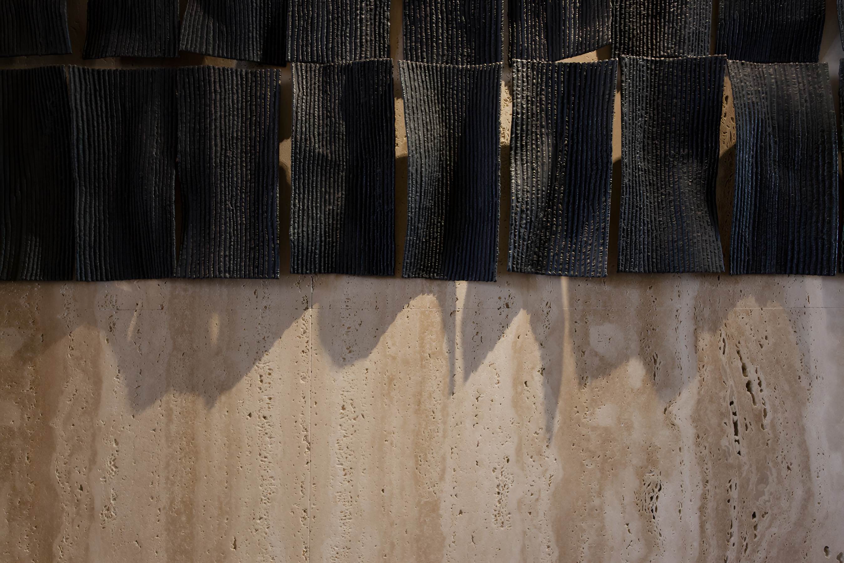 深色立體陶板藝術裝置懸掛於米色石材牆面上，展現建築材料與燈光投影的互動美感