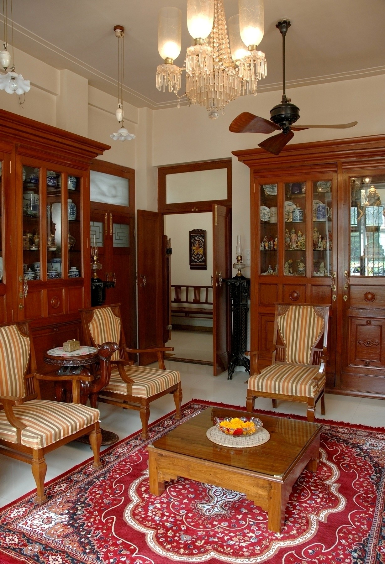 Traditional heritage living room with carved teak cabinetry, striped upholstered chairs, Persian rug and crystal chandelier.