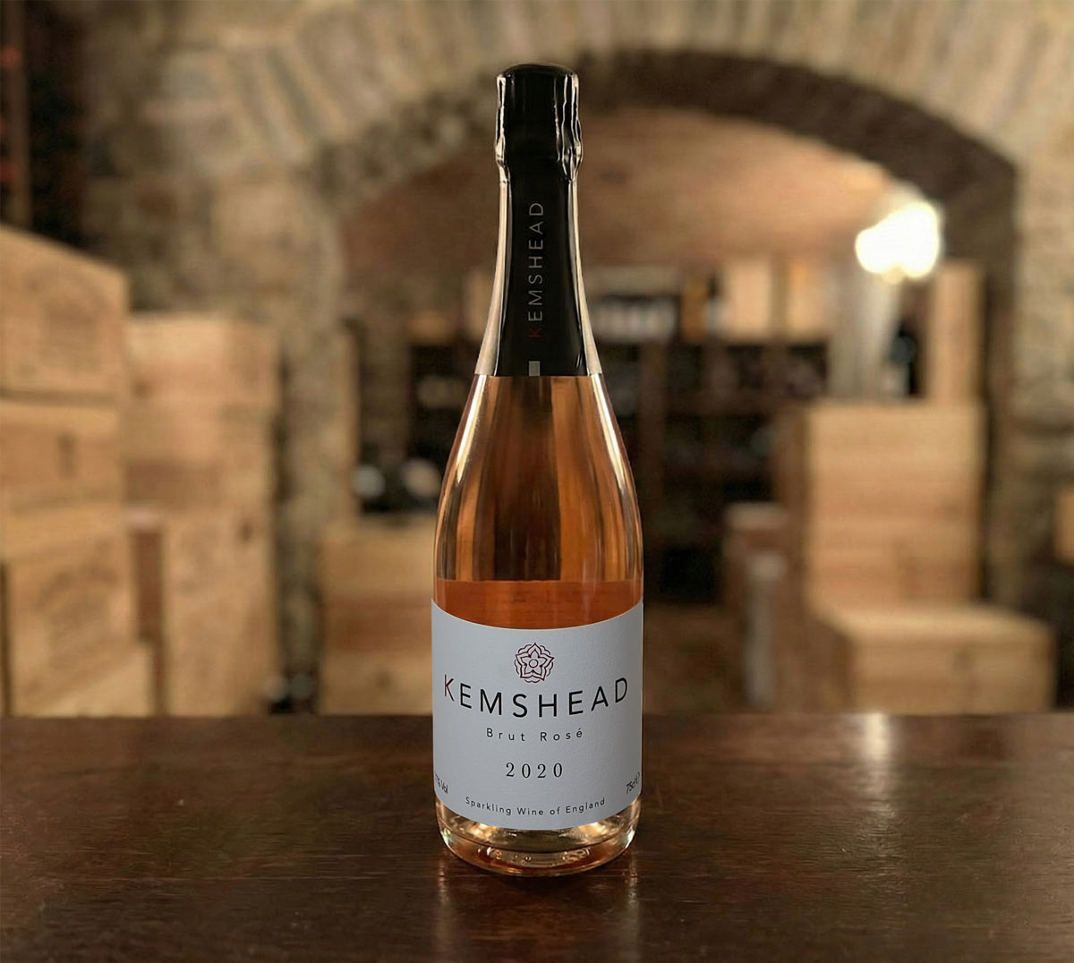 A bottle of Kemshead Brut Rosé 2020 sparkling wine standing on a dark wooden surface. The clear glass reveals a vibrant salmon-pink hue, and the bottle features a white label with the red rose logo and a black foil neck. The background is a blurred stone cellar with wooden wine barrels.
