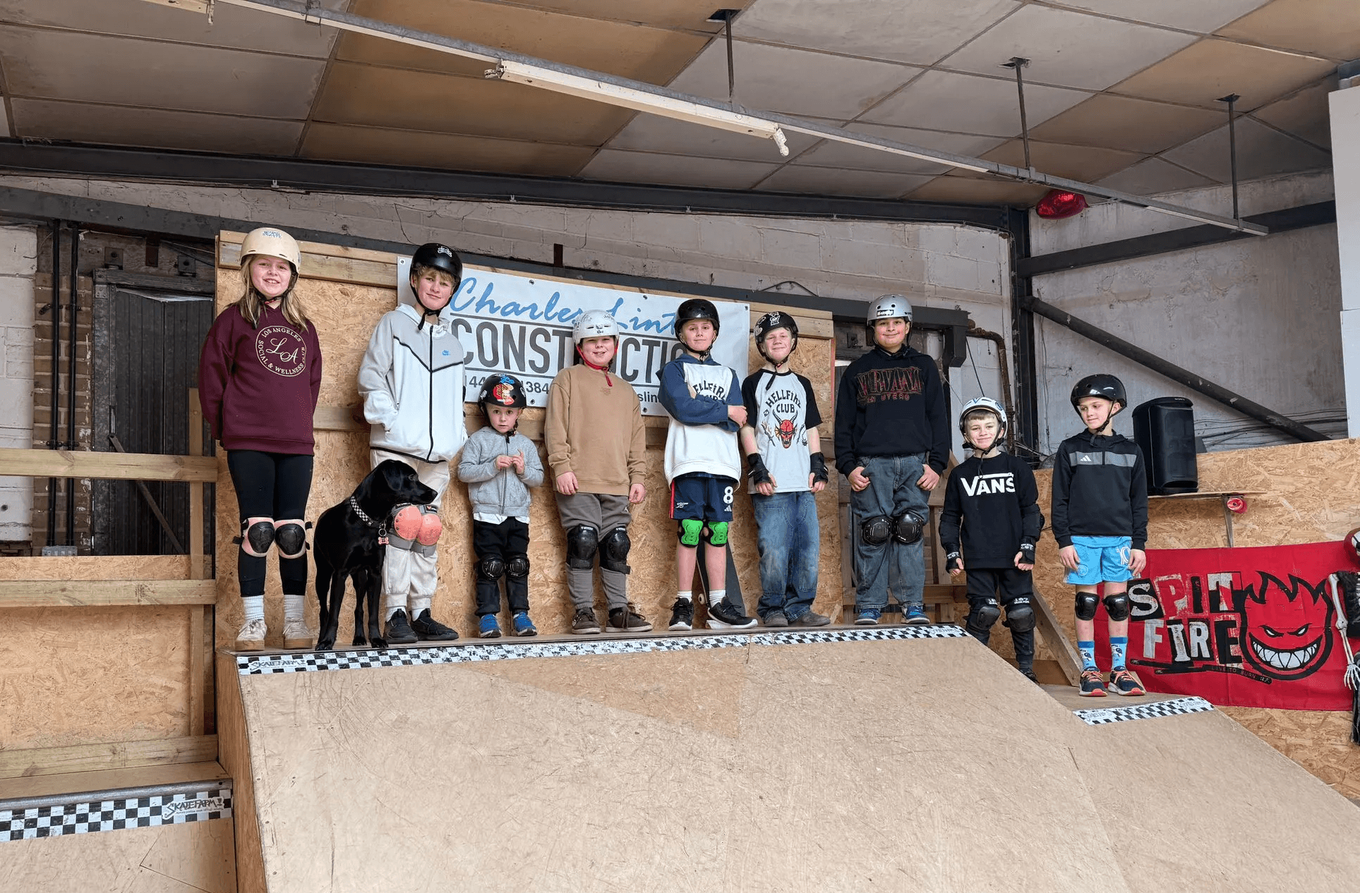 A group of six young people sitting together on a skate ramp, wearing helmets and skateboarding gear.