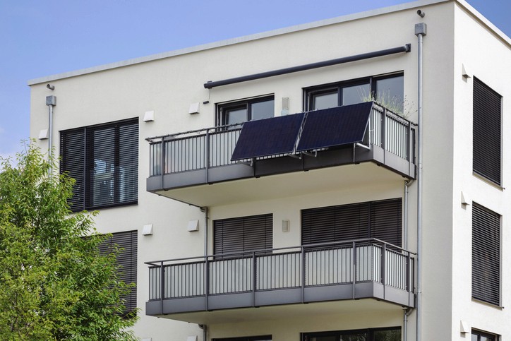 Modernes Gebäude mit Balkonen und Solarpaneelen, umgeben von Grünflächen unter einem klaren blauen Himmel.