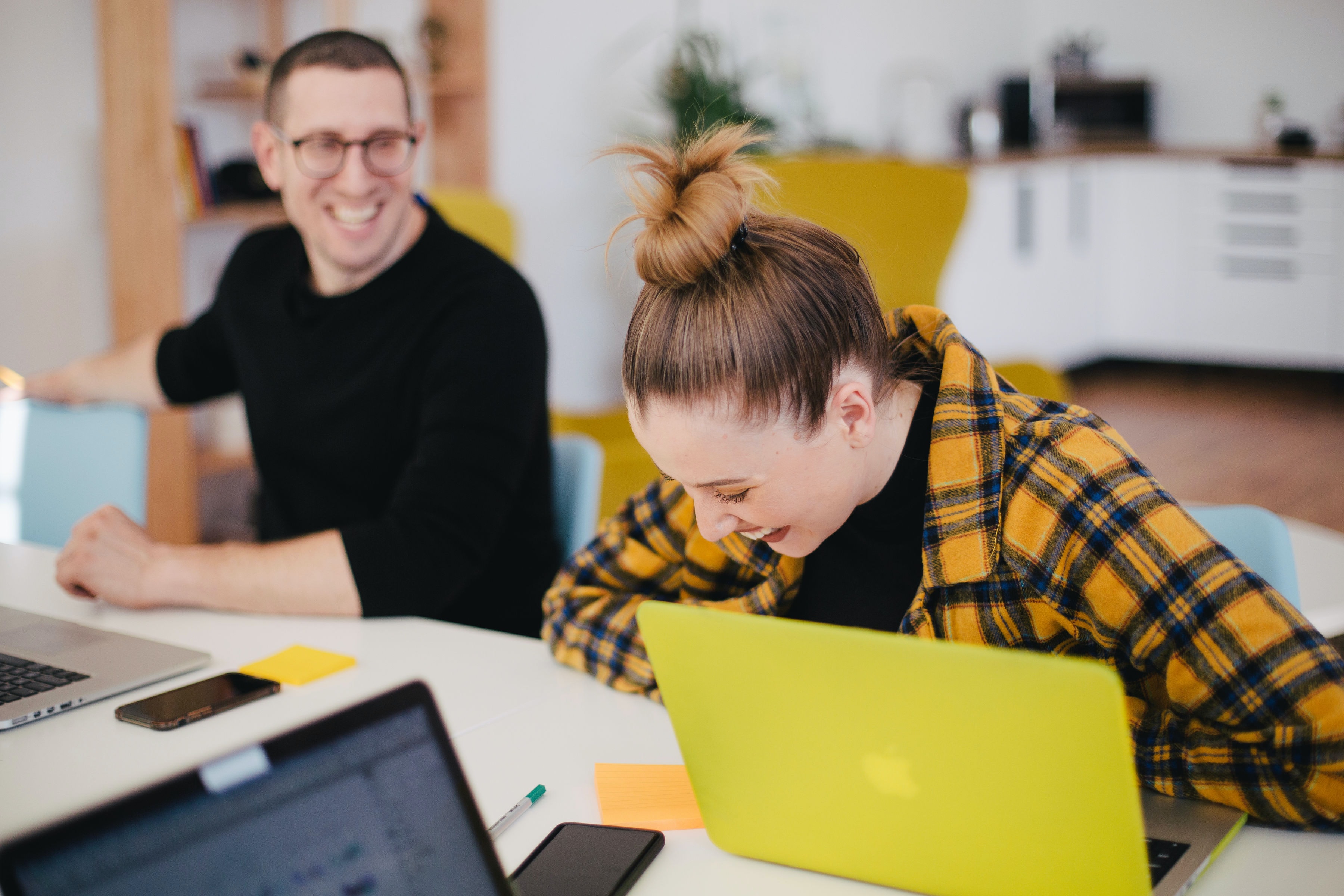 Two individuals are joyously laughing in the bright office space. They are both engaged via their respective laptops - one smartly-dressed individual dons a pair of glasses, while the other sports an elegant top bun hairstyle. Their optimistic laughter is resonating around the room as they participate in this session.