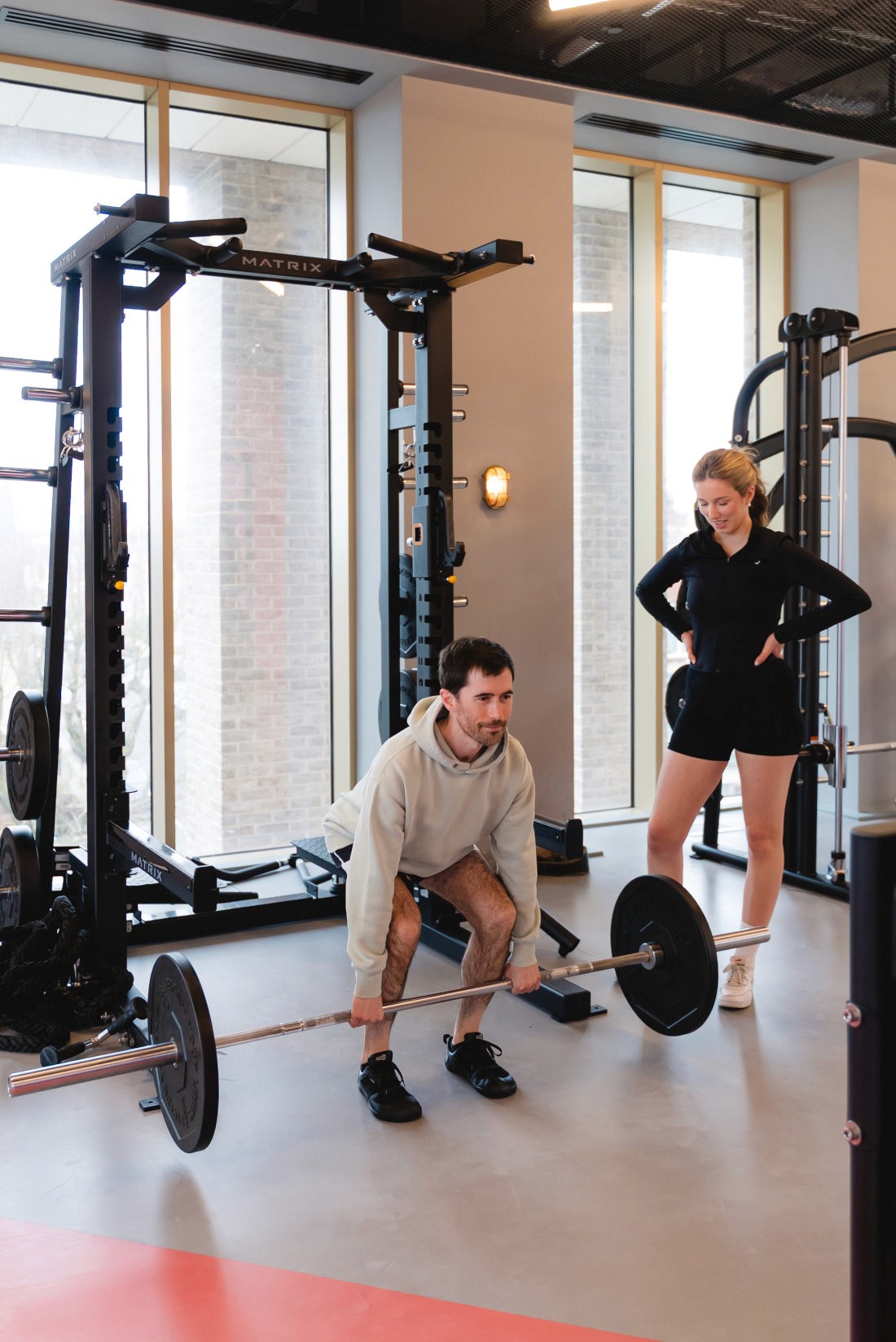 A man lifting a barbell in a gym while a woman watches from the side