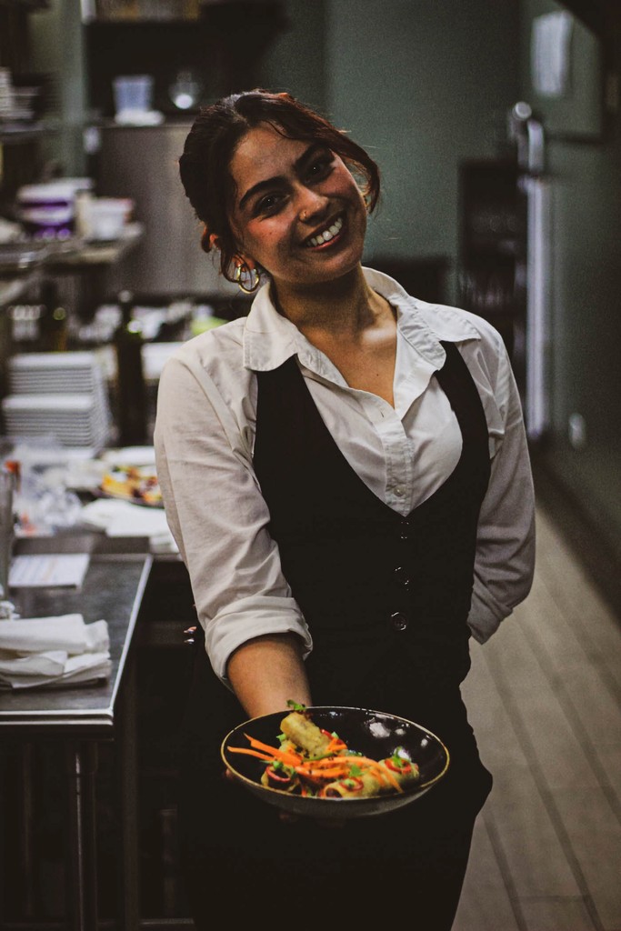 Smiling waitress in a restaurant kitchen, holding a plate of food.