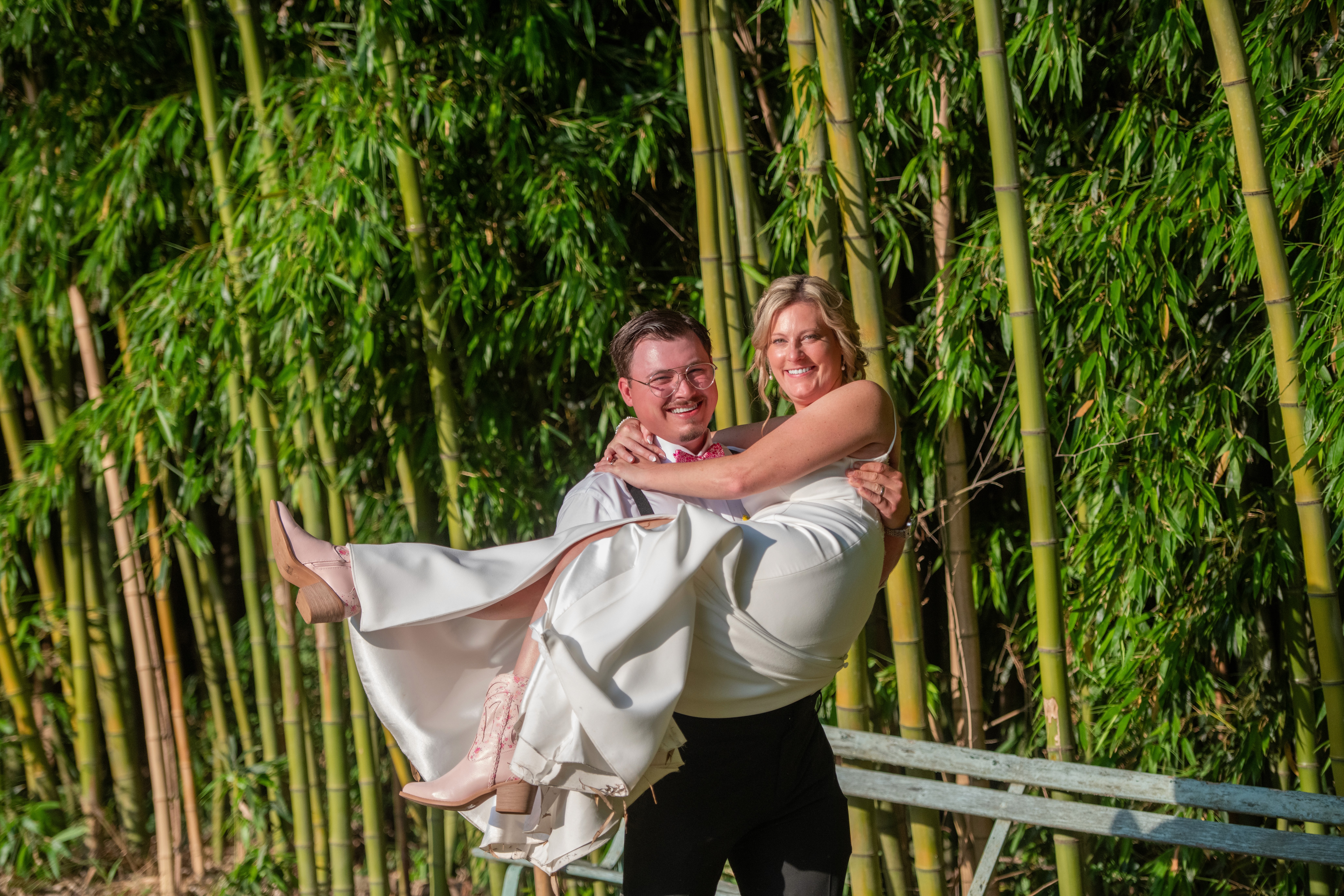 Happy groom holding his bride in his arms on their wedding day with a bamboo background.