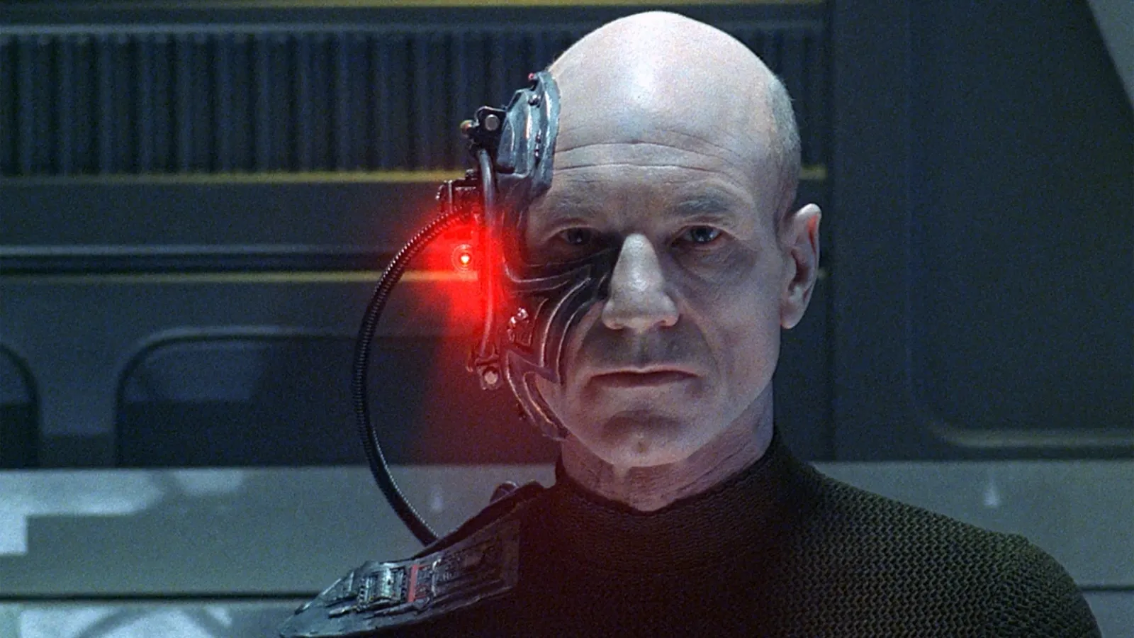 A bald man with a tense expression has a cybernetic implant on the left side of his face, emitting a red light, set against a futuristic, industrial backdrop.