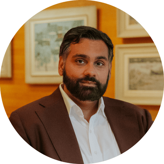 A person with dark hair and a beard, wearing a white shirt and a dark blazer, is standing in a warmly-lit room decorated with framed paintings on a wooden wall.